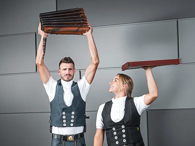 På bildet ser man to personer som hver holder en stabel taktekkingsmateriale i hendene. Personer til venstre holder en stabel takstein over hodet, mens personen til høyre holder en stabel aluminiumsplater over hodet. Begge har på seg tradisjonelle håndverkerklær med vest og hvit skjorte. Bakgrunnen er moderne og minimalistisk, med grå vegger. Begge personene virker konsentrerte og smiler.