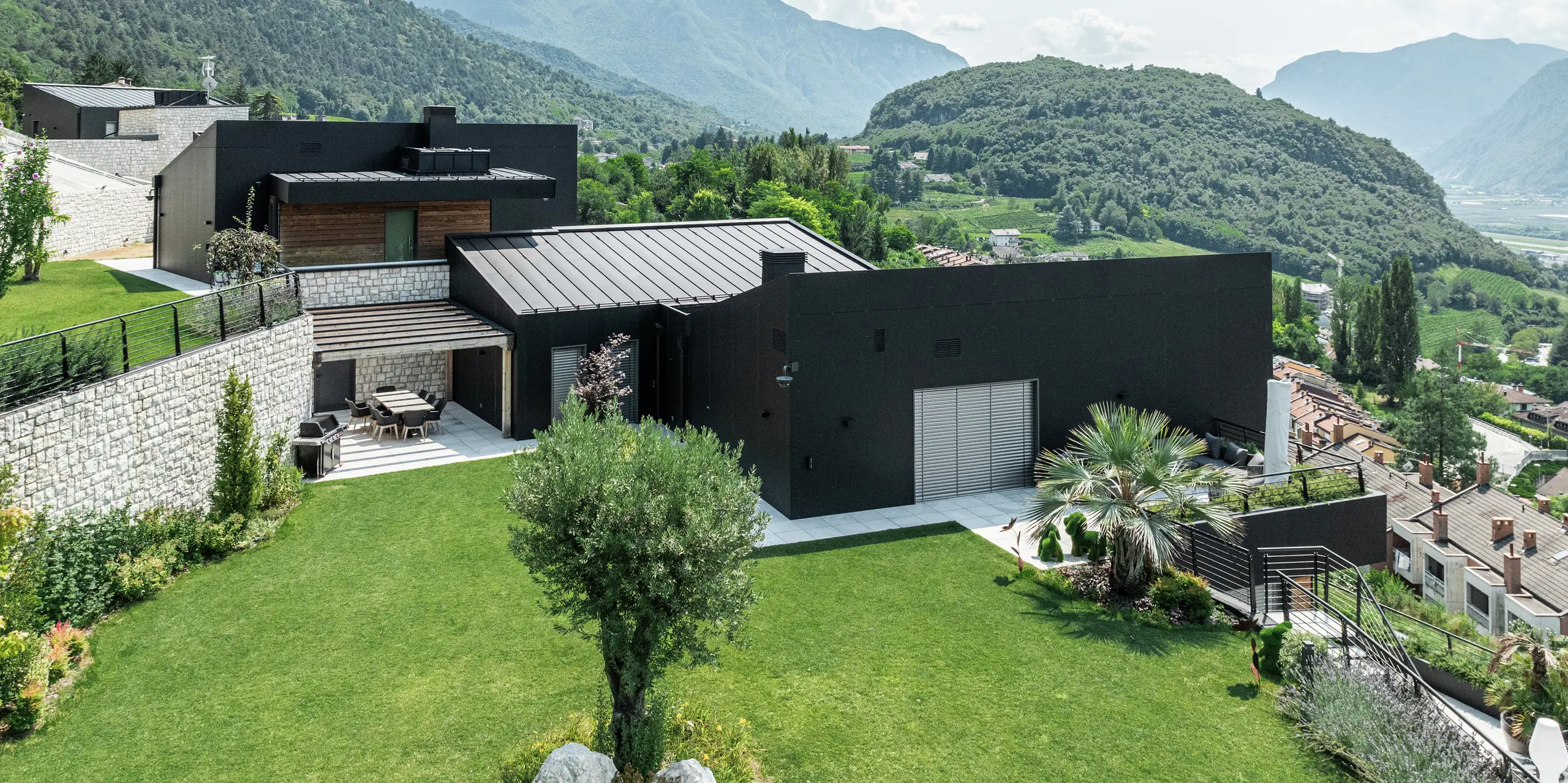 Moderne Wohnhausanlage in Hanglage mit atemberaubendem Blick auf die Trienter Berglandschaft. Die Fassade aus PREFA Aluminium Verbundplatten in P.10 Schwarz sorgt für eine elegante und witterungsbeständige Gebäudehülle. Das PREFALZ Dach in P.10 Schwarz rundet das minimalistische Design ab und bietet höchste Langlebigkeit. Eine gepflegte Gartenlandschaft mit Natursteinmauern fügt sich harmonisch in die Umgebung ein und unterstreicht den exklusiven Wohncharakter.