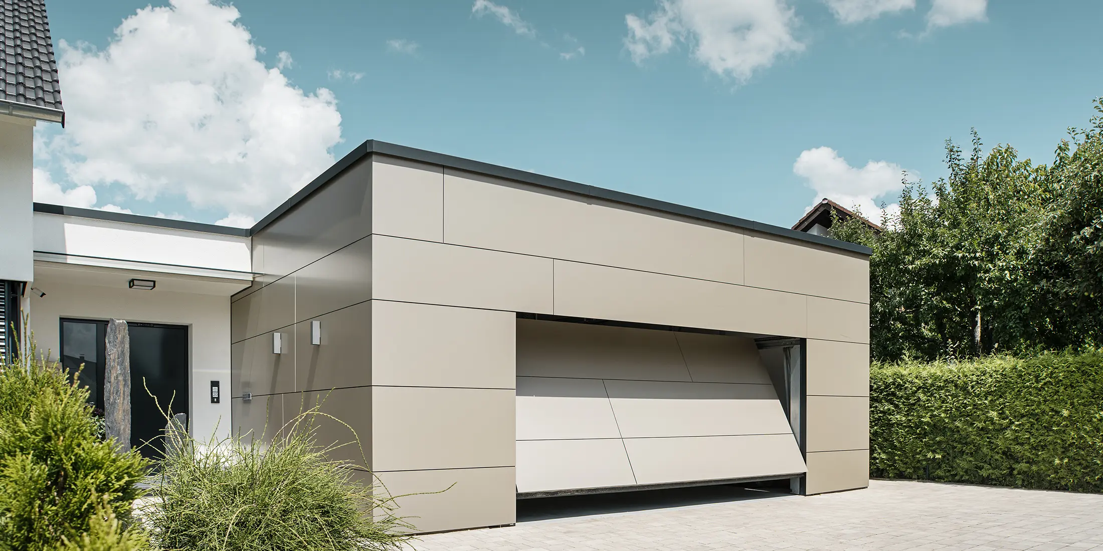 Gesamtansicht einer mit PREFABOND Aluminium Verbundplatten in Bronze verkleideten Garage. Das automatische Garagentor öffnet sich gerade und ist bereits ein Stück geöffnet. Direkt neben der Garage befindet sich der Hauseingang mit anthrazitfarbener Tür und weißer Putzfassade.