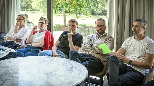 PREFARENZEN 2027 Jury-Meeting in einem hellen Seminarraum: Mehrere Architekten und Designexperten sitzen konzentriert in einer Reihe vor blauen Pinnwänden, auf denen Architekturprojekte mit PREFA Aluminium präsentiert werden. Große Fenster lassen Tageslicht herein und geben Blick auf einen grünen Garten frei. Auf den runden Tischen liegen Architekturbücher, Notizhefte und Stifte. Die Szene vermittelt intensive Diskussion, kreative Bewertung von Design und Funktion sowie die Inspiration, die PREFA Aluminiumprodukte durch langlebige, ästhetische Dächer und Fassaden in Architekturprojekten bieten.