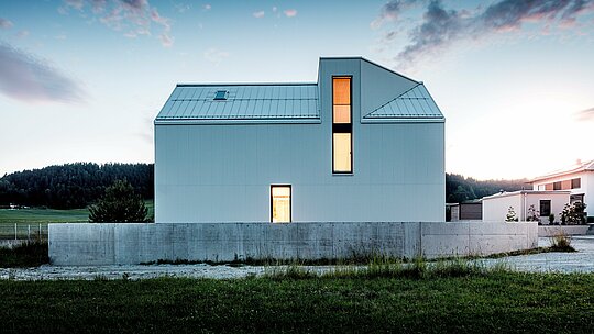 Das Bild zeigt das auffällige Einfamilienhaus in Freistadt, das durch seine klare Linienführung und die minimalistische Architektur besticht. Die reinweiße PREFALZ-Fassade aus gefaltetem Aluminium bildet eine markante, geometrische Struktur, die das Gebäude modern und zeitlos wirken lässt. Das steile Satteldach mit integriertem Gaubenfenster und die hohen Fensterflächen sorgen für viel Tageslicht und lassen den Innenraum offen und einladend erscheinen. Die Verwendung von PREFALZ-Aluminium garantiert nicht nur ästhetische Beständigkeit, sondern auch eine hervorragende Witterungsbeständigkeit und Langlebigkeit, was das Haus sowohl funktional als auch nachhaltig macht. Die klare Formensprache und die stilvollen Details tragen zu einer eleganten und zugleich praktischen Lösung für moderne Wohnarchitektur bei.