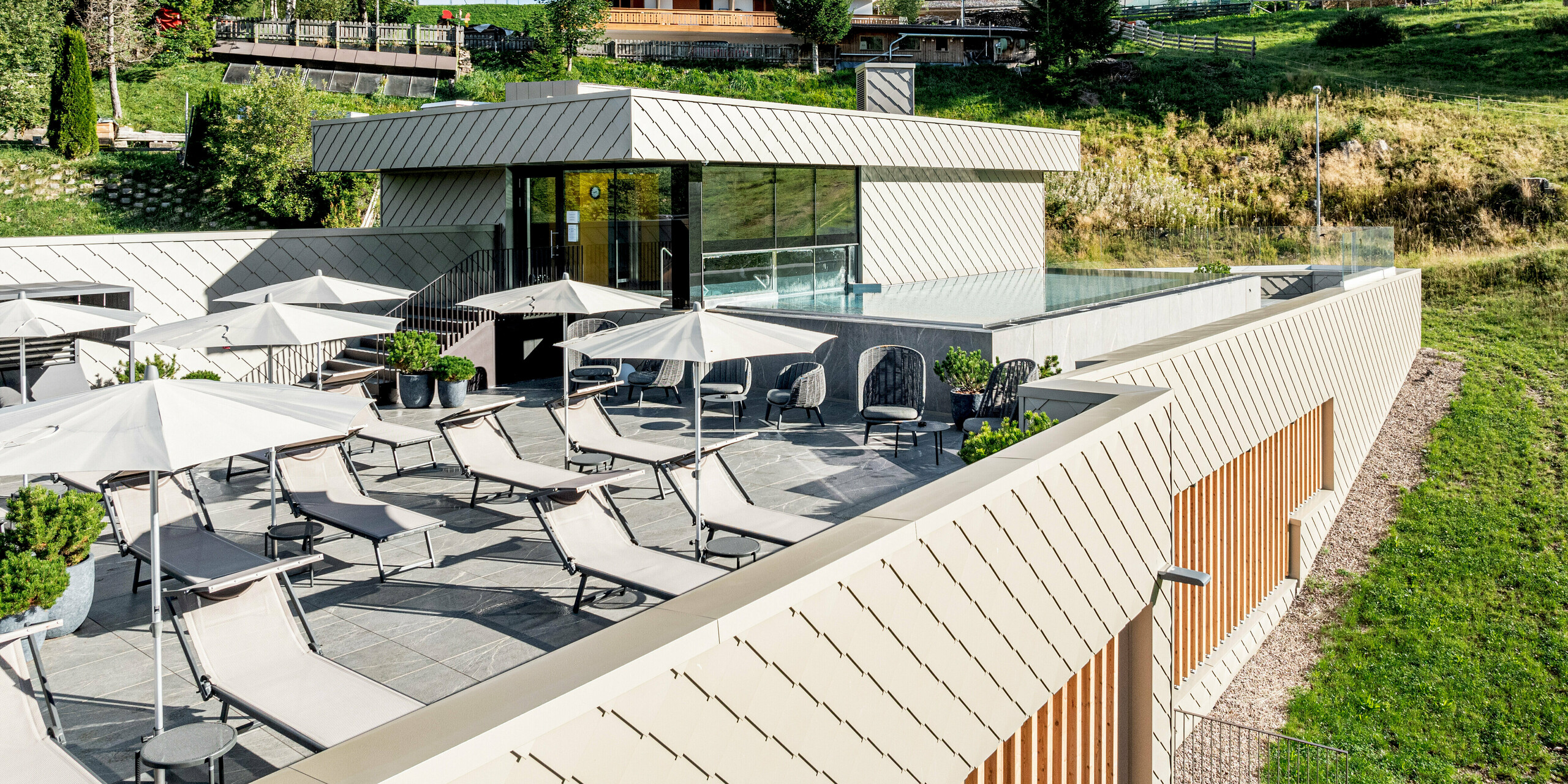 Terrassenbereich des Sporthotels Obereggen in Südtirol mit Outdoor-Pool, Liegen und Sonnenschirmen, eingebettet in die alpine Hanglandschaft. Die moderne Architektur wird durch die PREFA Aluminiumfassade mit Wandraute 29 × 29 in Bronze geprägt, die klare Linien und eine lebendige Oberflächenstruktur zeigt. Der warme Bronzeton harmoniert mit Holz und Natur. Die langlebigen, witterungsbeständigen und wartungsarmen PREFA Aluminiumprodukte bieten hohe Gestaltungsfreiheit und sind ideal für anspruchsvolle Hotelarchitektur im alpinen Raum.