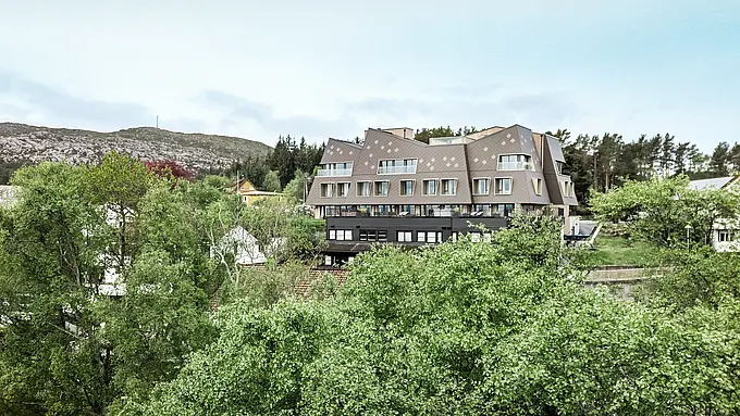 Utsikt på lang avstand fra boutiquehotellet Beckerwyc House i Bekkjarvik i Norge, som ligger i et grønt, kupert landskap. Den futuristisk utseende bygningen er kledd med PREFA-veggfliser 44 × 44 i valnøttbrun, som smelter harmonisk inn i de steinete omgivelsene. Hotellets uvanlige arkitektur skiller seg ut fra de omkringliggende tradisjonelle husene, samtidig som den passer godt inn i naturen takket være den jordnære fargeskalaen.