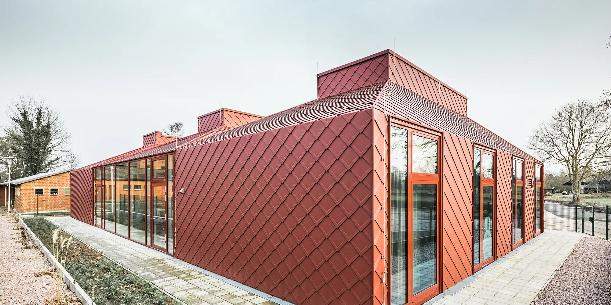 Ein modernes, einstöckiges Gebäude mit einer roten Fassade aus Aluminium, gedeckt mit PREFA Dach- und Wandrauten. Zu sehen sind große, raumhohe Fensterfronten sowie einer klaren, modernen Architektur, fotografiert bei Tageslicht in einer Frontalaufnahme.