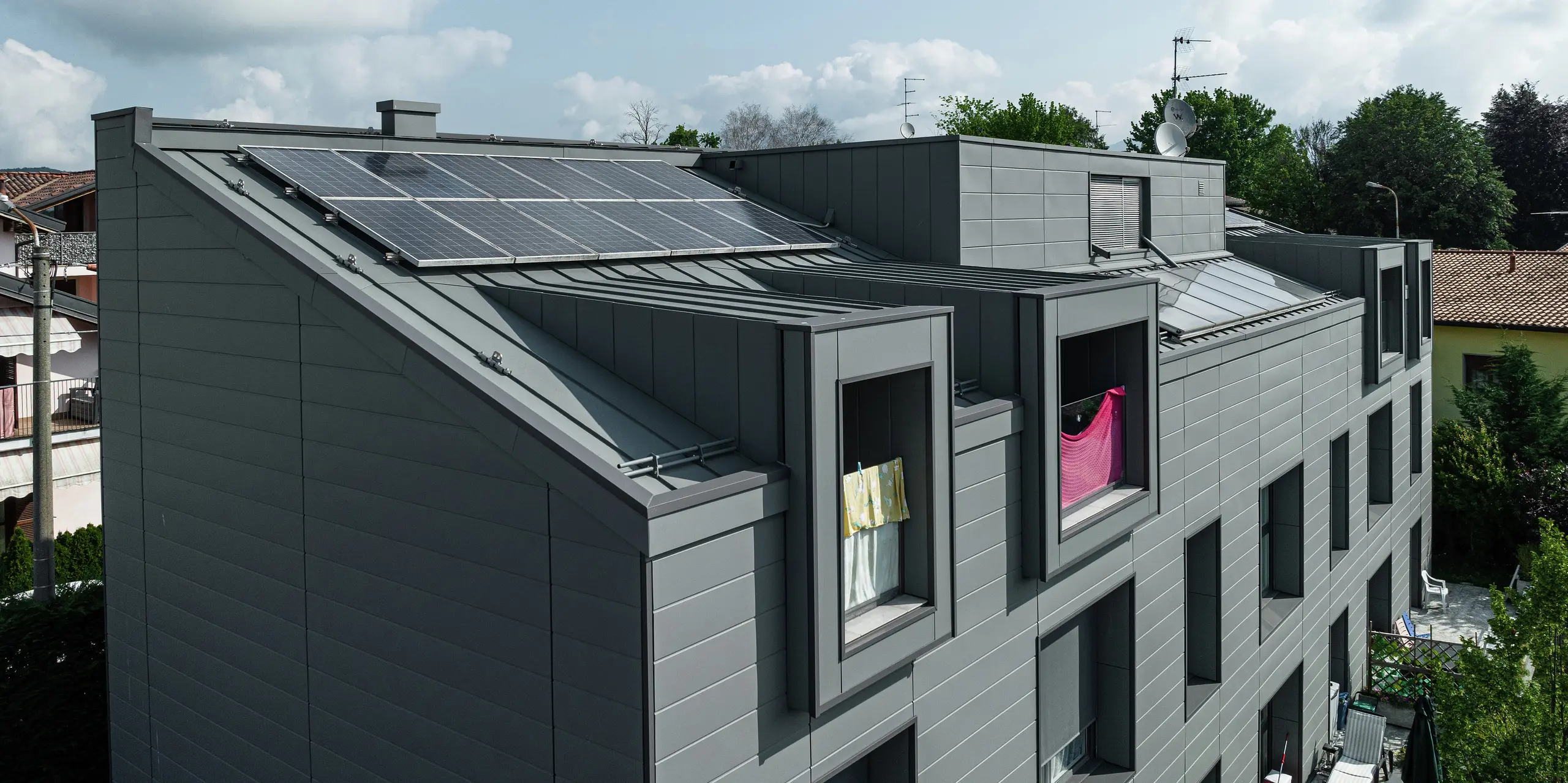 Vogelperspektive auf das Dach einer modernen Wohnanlage in Faloppio, Italien, mit einer robusten Stehfalzverkleidung aus Aluminium und einer nachhaltigen Photovoltaikanlage. Die PREFA Siding-Fassade und das Blechdach bieten nicht nur optische Highlights, sondern auch eine hervorragende Witterungsbeständigkeit und eine nachhaltige Energiequelle durch die PV-Module. Die Kombination aus funktionalen und ästhetischen PREFA Produkten sorgt für eine langlebige und energieeffiziente Lösung.