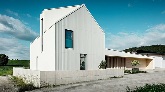 Das moderne Einfamilienhaus in Freistadt aus einer dynamischen Perspektive, die die klaren Linien und die auffällige Architektur betont. Die reinweiße PREFALZ-Aluminiumverkleidung zieht sich nahtlos über die gesamte Fassade und das Dach und verleiht dem Haus so eine moderne und zeitlose Ästhetik. Charakteristische Elemente des Entwurfs sind das steile Satteldach, die große Fensterfläche und die markante Gaube, während die vertikal verlaufenden Rillen der Fassade dem Gebäude eine interessante Textur und Tiefe verleihen. Der Kontrast zur natürlichen Umgebung sowie die eleganten Holzelemente im Eingangsbereich und am Garagentor ergänzen das Design. PREFALZ sorgt für eine besonders langlebige, pflegeleichte und witterungsbeständige Oberfläche. Dadurch wird das Haus nicht nur ästhetisch ansprechend, sondern auch funktional und nachhaltig.