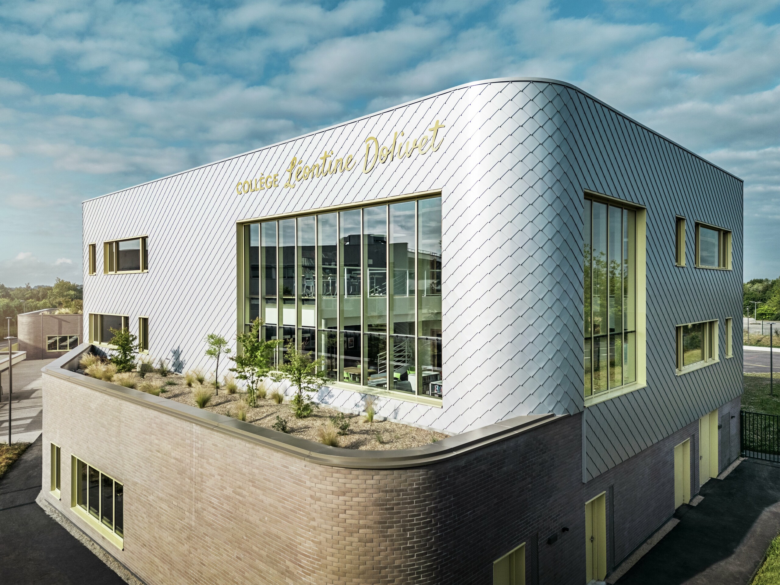 Das Foto zeigt eine markante Ecke des Collège Léontine Dolivet in Cesson-Sévigné. Die Fassade besteht aus silbermetallischen PREFA-Wandelementen, die diagonal angeordnet sind und das Licht reflektieren. Besonders auffällig ist die geschwungene Form des Obergeschosses, die dem Gebäude eine sanfte und moderne Anmutung verleiht. Große Glasflächen geben den Blick ins Innere frei, während gelbe Fensterrahmen farbliche Akzente setzen. Im Vordergrund befindet sich eine bepflanzte Fläche mit Kies und Sträuchern, die die Architektur harmonisch ergänzt. Der leicht bewölkte Himmel hebt die Metalloberfläche zusätzlich hervor.
