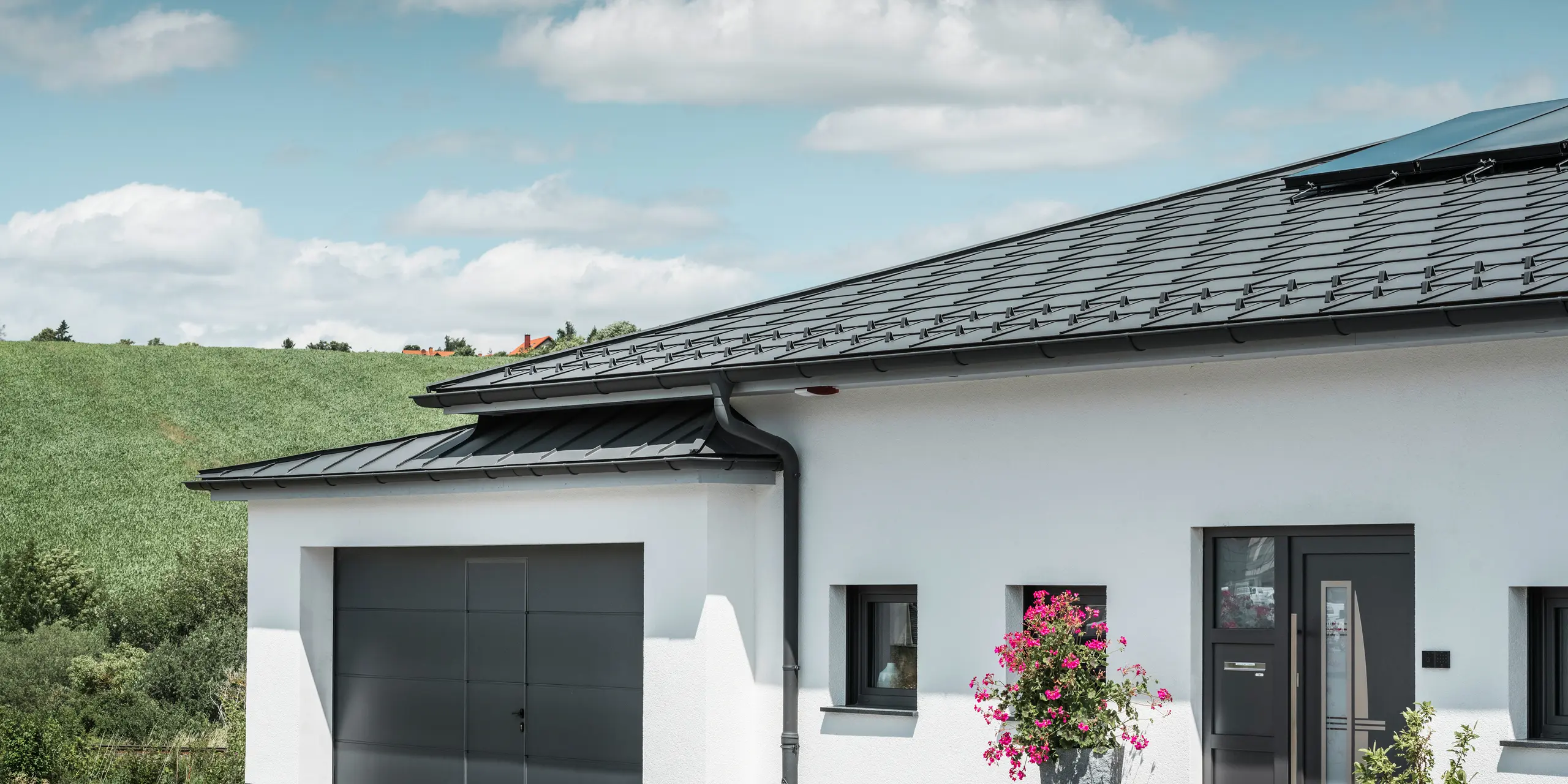 Bildet viser framsiden av en enebolig i Ilmenau, Thüringen, med et elegant PREFA-tak i P.10 mørk grå. Takplaten R.16 gir huset et moderne utseende, mens det integrerte PREFA takavløpssystemet med sin mørk grå takrenne og nedløpsrør garanterer effektiv avrenning av regnvann. Garasjen, utstyrt med mørk grå PREFALZ, passer harmonisk inn i den generelle estetikken og understreker kvaliteten og stilen til PREFA aluminiumsprodukter.