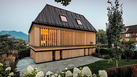 Das moderne Badehaus des Hotels Hirschen in Schwarzenberg verfügt über ein preisgekröntes PREFA Stehfalzdach in der Farbe P.10 Nussbraun. Das ikonische Walmdach mit 60-Grad-Neigung und unkonventionell gesetzten Stehfalzen ist ein architektonisches Highlight, das Tradition und Innovation verbindet. Die Fassade aus vertikalen Holzlamellen verleiht dem Gebäude eine warme, natürliche Ausstrahlung und fügt sich harmonisch in den denkmalgeschützten Ortskern ein. PREFALZ überzeugt durch Langlebigkeit, Formfreiheit und exzellente Verarbeitbarkeit, auch bei komplexen Dachformen. Ein preisgekröntes Beispiel für innovative Architektur mit hochwertigen PREFA-Produkten.
