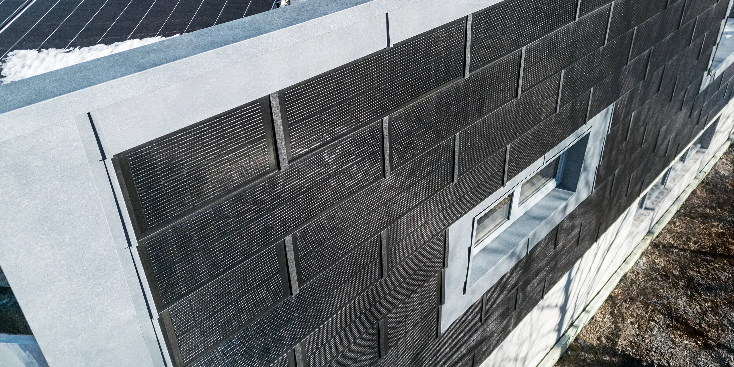Die Luftaufnahme zeigt die Fassade der RÜMA-Blechwerkstatt in Vorarlberg mit grauen FX.12-Fassadenpaneelen und innovativen PREFA-Solardachpaneelen mit integrierten Photovoltaikmodulen. Im Vordergrund ist ein teilweise schneebedecktes Flachdach mit weiteren Photovoltaikmodulen zu sehen.