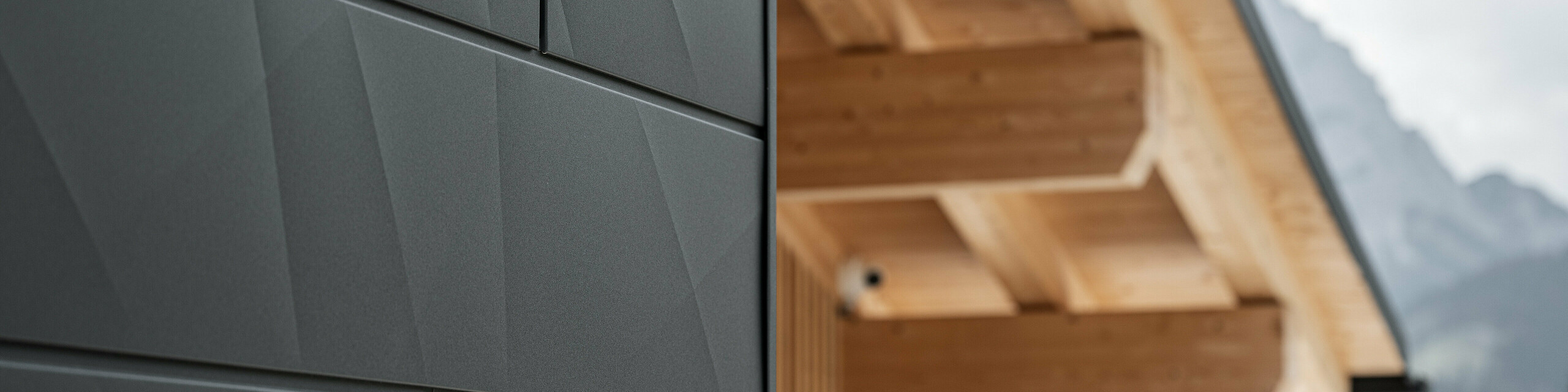 Nærbilde av Siding.X fasadeplater i P.10 svart fra PREFA på Casa Hoffe i Sappada, Italia. Fasadesystemet i aluminium er dyktig montert i et moderne, abstrakt design som danner en tiltalende kontrast til de synlige treelementene i takkonstruksjonen. Den innovative fasadeløsningen passer perfekt inn i det alpine miljøet og står for en kombinasjon av funksjonalitet, holdbarhet og estetisk byggekultur.