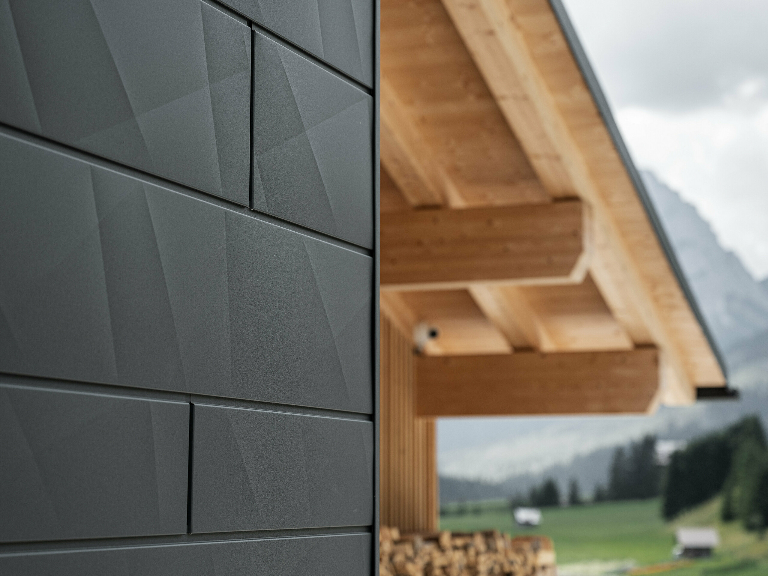 Nærbilde av Siding.X fasadepaneler i P.10 svart fra PREFA på Casa Hoffe i Sappada, Italia. Fasadesystemet i aluminium er montert i et moderne, abstrakt design som skaper en vakker kontrast til de synlige treelementene i takkonstruksjonen. Den innovative fasadeløsningen glir sømløst inn i det alpine miljøet og representerer en kombinasjon av funksjonalitet, holdbarhet og estetisk byggeskikk.