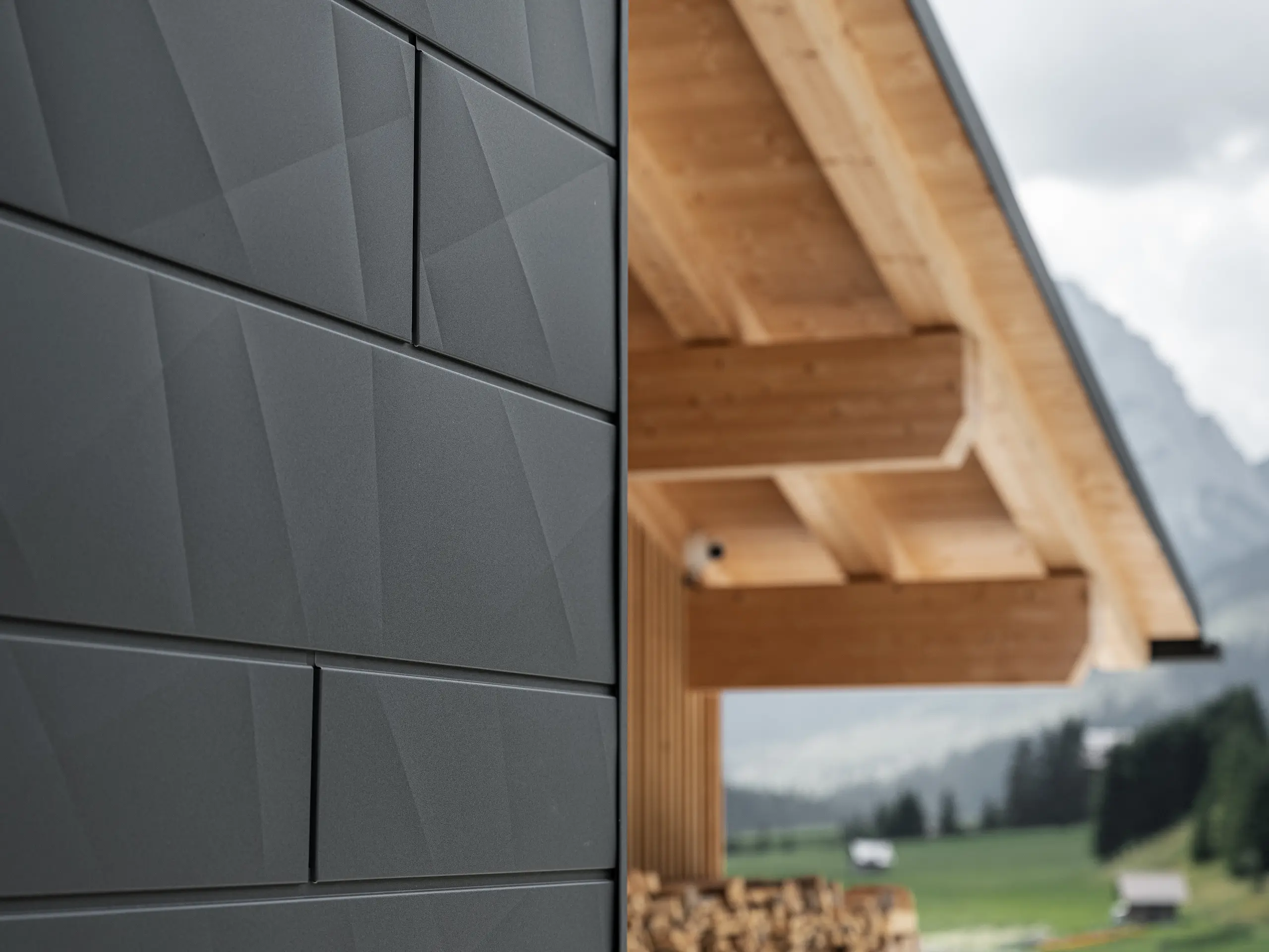 Nærbilde av Siding.X fasadepaneler i P.10 svart fra PREFA på Casa Hoffe i Sappada, Italia. Fasadesystemet i aluminium er montert i et moderne, abstrakt design som skaper en vakker kontrast til de synlige treelementene i takkonstruksjonen. Den innovative fasadeløsningen glir sømløst inn i det alpine miljøet og representerer en kombinasjon av funksjonalitet, holdbarhet og estetisk byggeskikk.