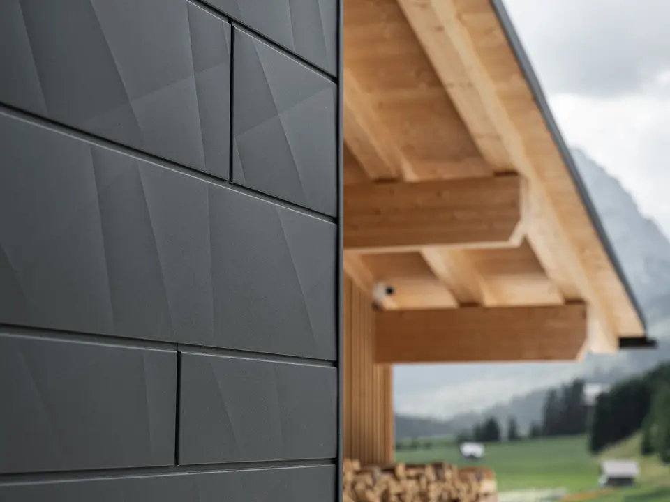 Nærbilde av Siding.X fasadepaneler i P.10 svart fra PREFA på Casa Hoffe i Sappada, Italia. Fasadesystemet i aluminium er montert i et moderne, abstrakt design som skaper en vakker kontrast til de synlige treelementene i takkonstruksjonen. Den innovative fasadeløsningen glir sømløst inn i det alpine miljøet og representerer en kombinasjon av funksjonalitet, holdbarhet og estetisk byggeskikk.