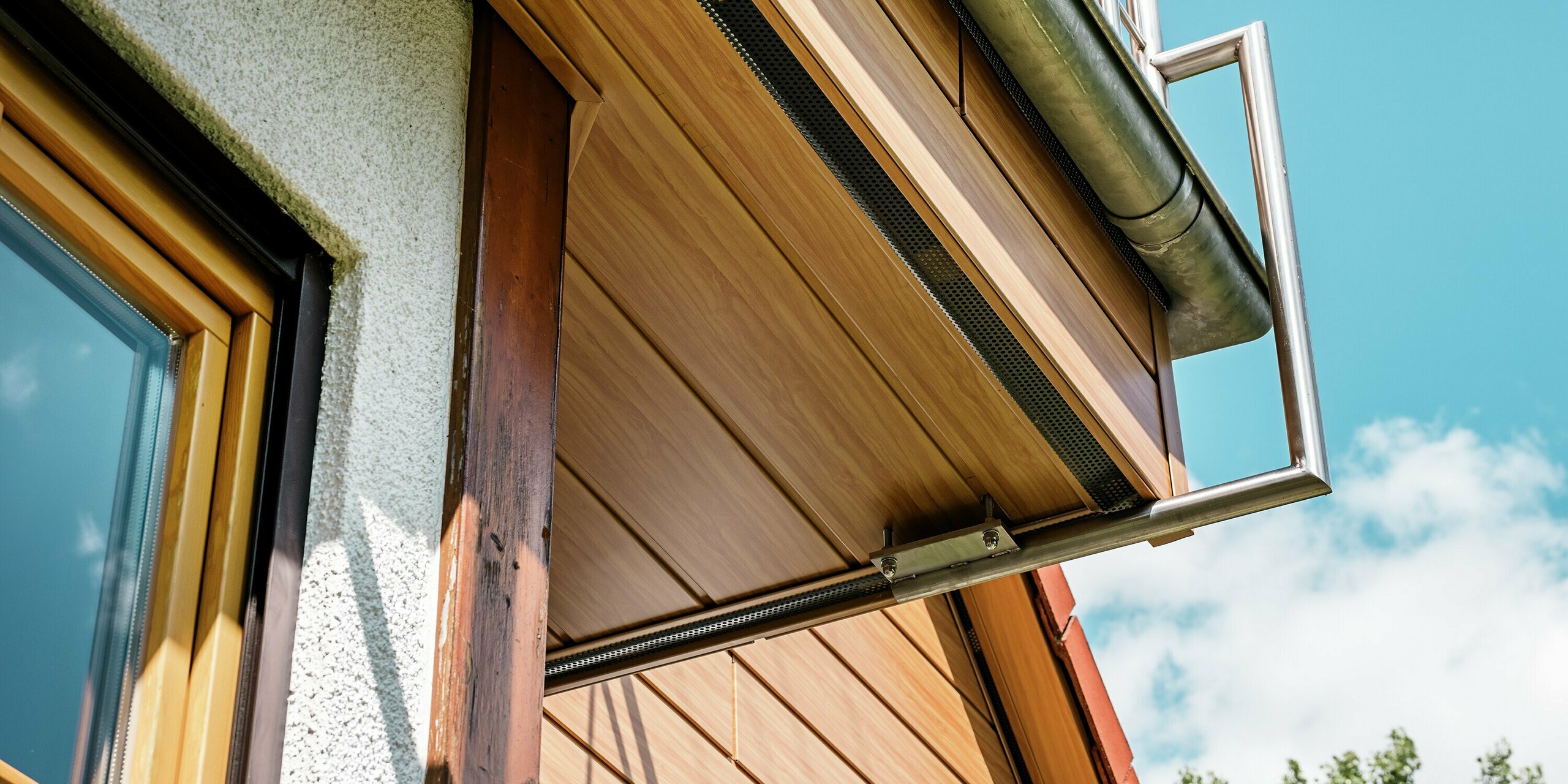 Das Bild zeigt eine dynamische Nahaufnahme des sanierten Giebels eines Hauses in Blieskastel, verkleidet mit PREFA Sidings in Eiche Natur. Aus der Froschperspektive wirken die Holzoptik-Paneele besonders authentisch und hochwertig, während das filigrane Balkon­geländer aus Metall elegant darüber schwebt. Die warmen Farbtöne der Aluminiumfassade harmonieren mit dem sonnigen Himmel und dem üppigen Grün im Vordergrund. Dank der witterungsbeständigen, formstabilen PREFA Oberfläche bleibt die natürliche Optik dauerhaft erhalten – ein stilvolles Detail, das das gesamte Gebäude aufwertet.