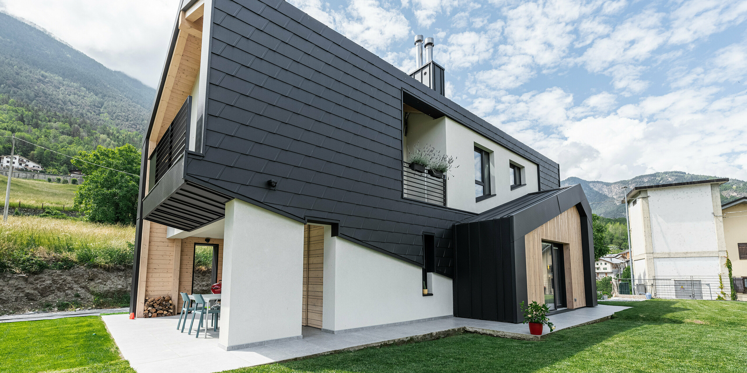 Blick vom Garten auf ein modernes Wohnhaus mit dem klingenden Namen „Zaffhaus“. Das Einfamilienhaus mit einer Fassade aus schwarzen PREFA Wandschindeln, zeichnet sich durch seine markante Architektur aus. Das Objekt besticht durch den eindrucksvollen Kontrast aus dunklen Aluminiumschindeln, hellen Holzelementen und weißen Putzflächen. Diese spannende Kombination schafft ein ästhetisches Gesamtbild, das durch die umliegende grüne Wiese und die bergige Landschaft im Hintergrund abgerundet wird.