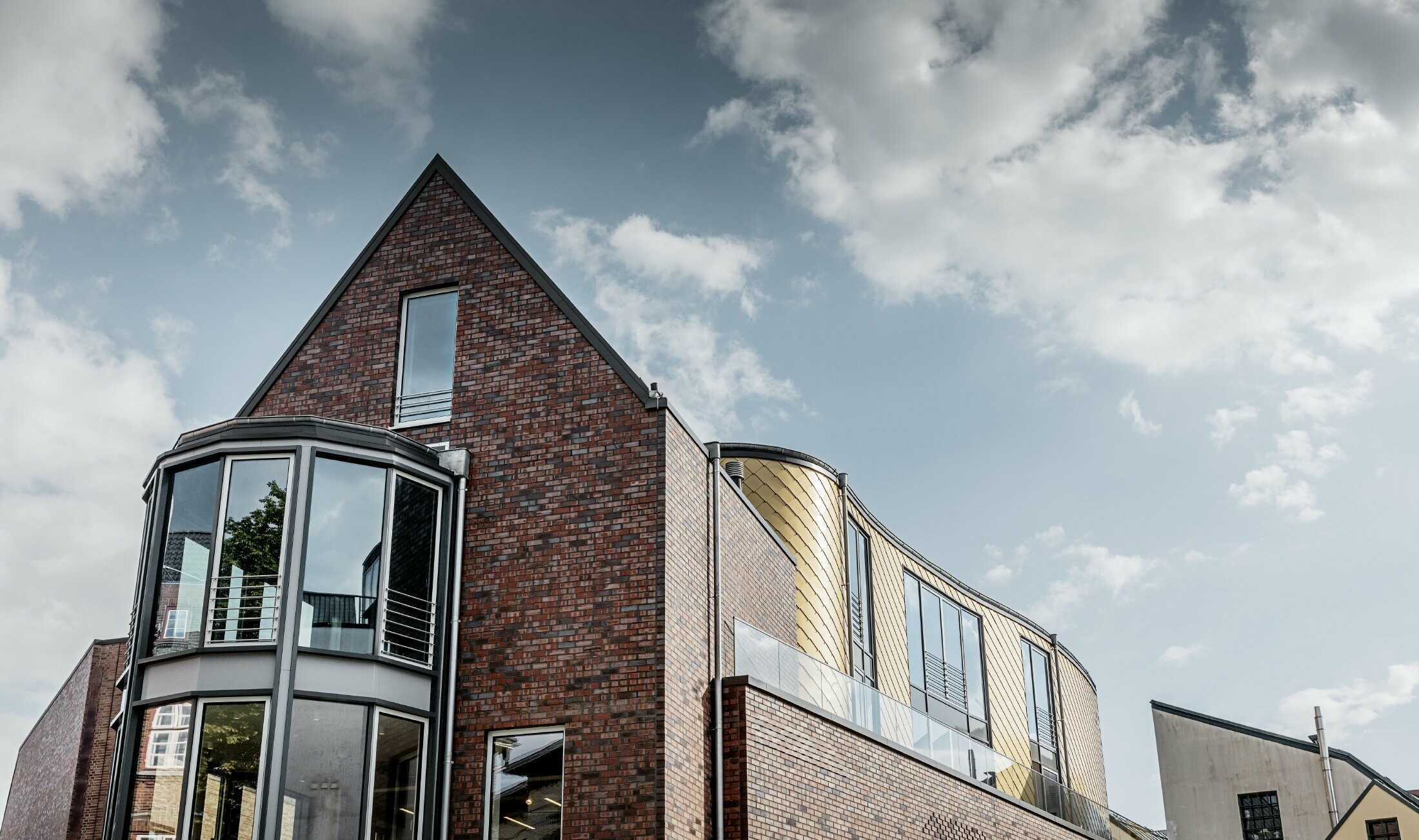 Forretningsbygg med mursteinsfasade og sjenerøse glassflater. En del av den buede fasaden er kledd med den gylne veggromben av aluminium fra PREFA.