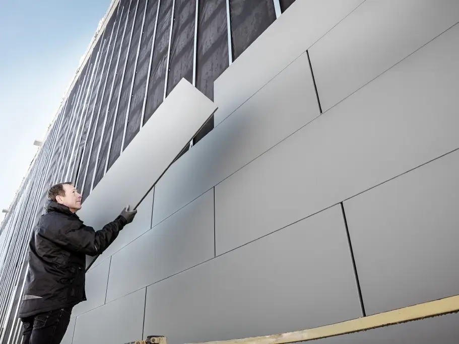 Bilder av en delvis ferdigstilt fasade som er kledd med PREFA Siding i bredde 500. Blikkenslageren monterer et nytt fasadepanel. Bildet er tatt nedenfra. 