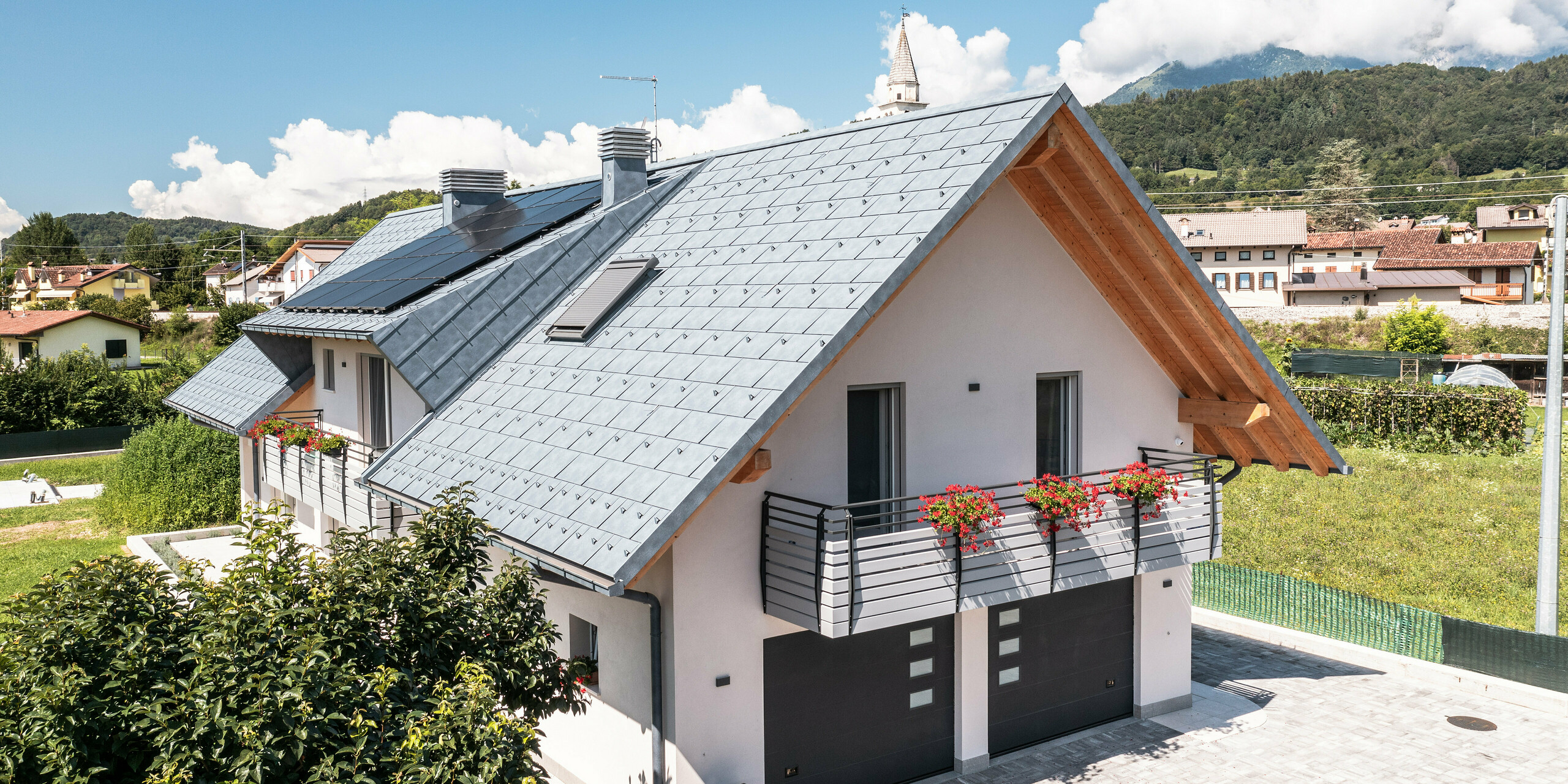 Modernes Einfamilienhaus in Bellun, Italien, geprägt durch das PREFA Dachsystem in der Farbe Anthrazit in Form der Dachplatte R.16, die optisch zu den sichtbaren Holzbalken des Giebels passt. Ein sorgfältig gestalteter Außenbereich mit blühenden Balkonkästen und einer gepflegten Hecke verleiht dem Anwesen in einem ruhigen Dorf vor der Kulisse der Dolomiten ein einladendes Flair. Unter dem Dach befinden sich zwei Garageneinfahrten. Das Garagentor ist farblich auf die Aluminiumelemente abgestimmt und verleiht der Immobilie ein harmonisches Gesamtbild.