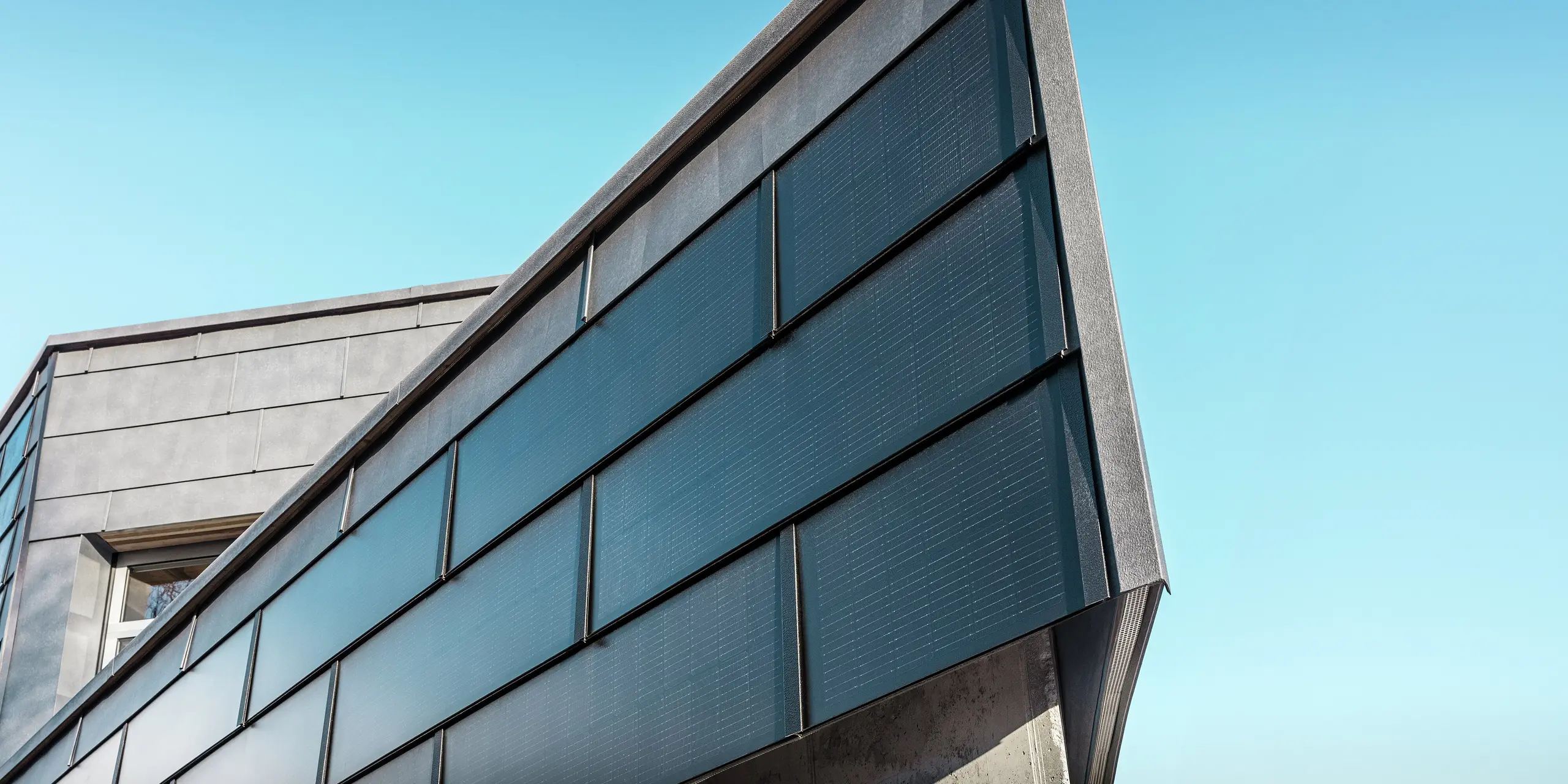 Moderne Geschäftsfassade der RÜMA Blechwerkstatt in Vorarlberg mit PREFA Solar-Aluminium-Dachpaneelen und integrierten Photovoltaikmodulen, kombiniert mit PREFA Fassadenpaneelen FX.12 für nachhaltige Energieerzeugung und dynamische Fassadengestaltung. Die Aufnahme zeigt eine Nahaufnahme einer Gebäudeecke. Im Hintergrund ist ein klarer blauer Himmel zu sehen.
