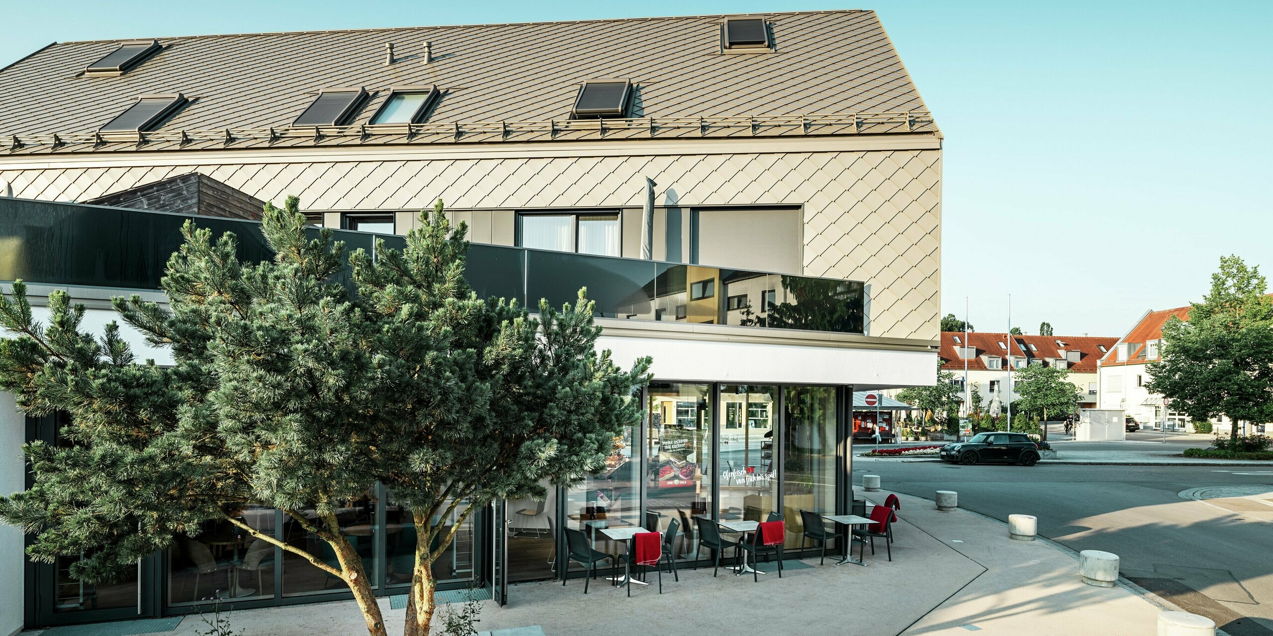 Modernes Mehrfamilienhaus mit markanter Fassadengestaltung in Leitershofen, Deutschland. Das Gebäude ist mit widerstandsfähigen PREFA Dach- und Wandrauten 29 × 29 in der Farbe Bronze eingedeckt, die eine stilvolle und zeitlose Optik erzeugen. Die Kombination aus klaren Linien, großflächigen Fenstern und einer hellen Putzfassade im Erdgeschoss unterstreicht die hochwertige Architektur. Die Dach- und Fassadenelemente aus Aluminium bieten neben der ästhetischen Eleganz auch eine hohe Witterungsbeständigkeit und Langlebigkeit.