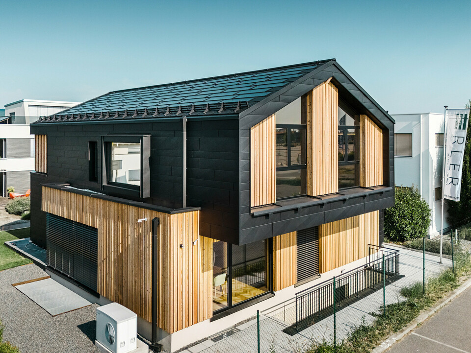 framside av mønsterhuset Autark i prefabrikksenteret Blaue Lagune, Vösendorf (AT). Taket er dekket med svarte PREFA solcelle takplater og takplate R.16 i høy kvalitet P.10-lakkering. Fasade består av en kombinasjon av vertikale treelementer og PREFA Siding.X i P.10 svart, noe som gir bygningen et moderne og bærekraftig preg. På høyre side av bildet vaier flagget til Erler Bau GmbH.