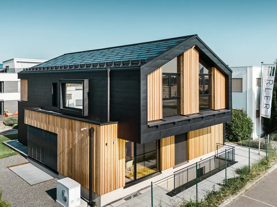 framside av mønsterhuset Autark i prefabrikksenteret Blaue Lagune, Vösendorf (AT). Taket er dekket med svarte PREFA solcelle takplater og takplate R.16 i høy kvalitet P.10-lakkering. Fasade består av en kombinasjon av vertikale treelementer og PREFA Siding.X i P.10 svart, noe som gir bygningen et moderne og bærekraftig preg. På høyre side av bildet vaier flagget til Erler Bau GmbH.
