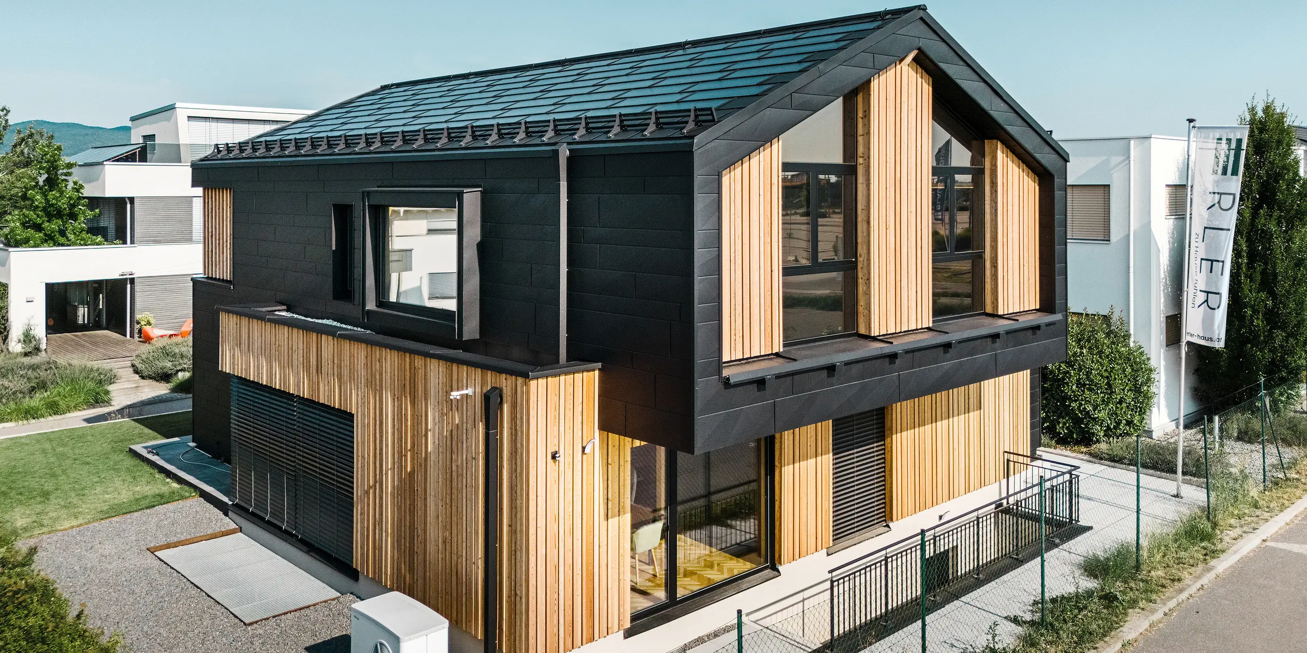 framside av mønsterhuset Autark i prefabrikksenteret Blaue Lagune, Vösendorf (AT). Taket er dekket med svarte PREFA solcelle takplater og takplate R.16 i høy kvalitet P.10-lakkering. Fasade består av en kombinasjon av vertikale treelementer og PREFA Siding.X i P.10 svart, noe som gir bygningen et moderne og bærekraftig preg. På høyre side av bildet vaier flagget til Erler Bau GmbH.