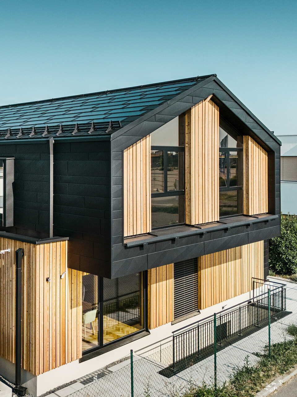 framside av mønsterhuset Autark i prefabrikksenteret Blaue Lagune, Vösendorf (AT). Taket er dekket med svarte PREFA solcelle takplater og takplate R.16 i høy kvalitet P.10-lakkering. Fasade består av en kombinasjon av vertikale treelementer og PREFA Siding.X i P.10 svart, noe som gir bygningen et moderne og bærekraftig preg. På høyre side av bildet vaier flagget til Erler Bau GmbH.