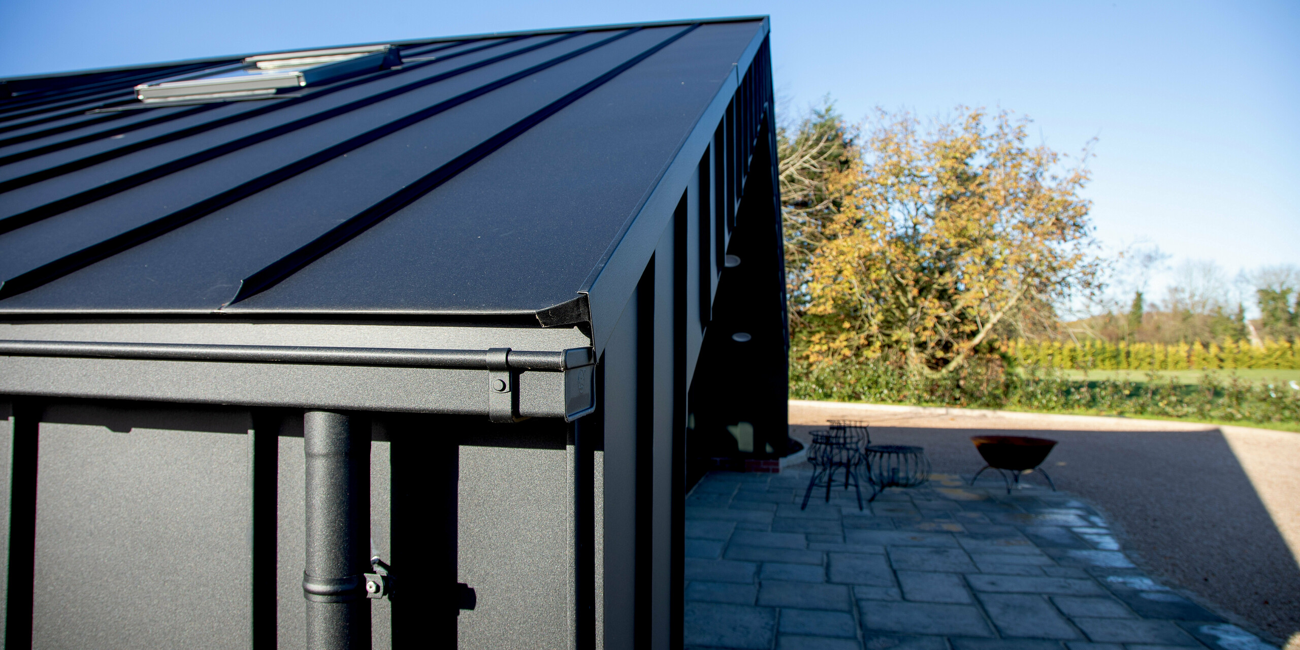 Detailansicht der modernen PREFA Gebäudehülle mit PREFALZ in P.10 Schwarz. Die klaren Stehfalzlinien und die präzise ausgeführte Aluminium-Traufe formen eine markante, elegante Architektur. Die witterungsbeständige Aluminiumoberfläche sorgt für hohe Stabilität und schützt das Freizeitgebäude dauerhaft, während der Außenbereich harmonisch in die moderne Gestaltung übergeht.