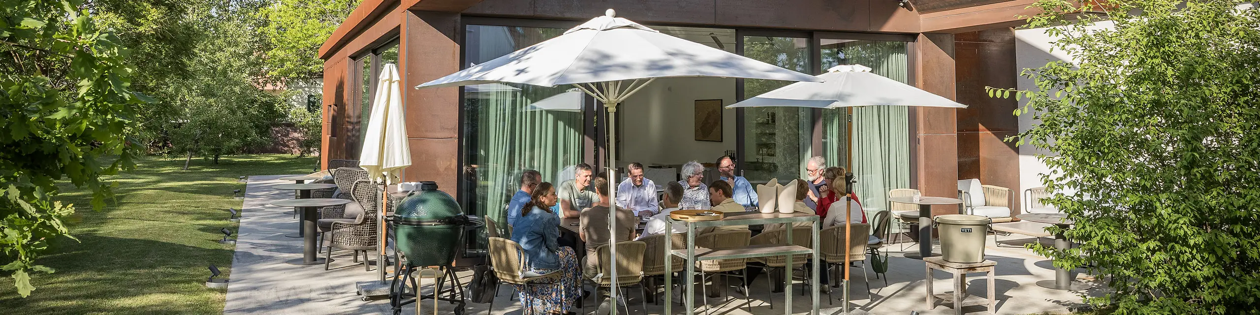 PREFARENZEN 2027 Expertendialog auf der Terrasse einer modernen Winzerresidenz: Jury-Mitglieder sitzen um einen großen Tisch, diskutieren intensiv über Architekturprojekte und blättern in Architekturbüchern. Die Szene wird von natürlichem Licht und grünem Garten umrahmt, während PREFA Aluminiumprodukte an Dach und Fassade des Gebäudes sichtbar sind. Das Bild vermittelt kreative Auseinandersetzung, konzentrierte Analyse und lebendigen Austausch. PREFA Dächer und Fassaden stehen für innovative, langlebige und ästhetisch anspruchsvolle Lösungen, die Design, Funktion und Inspiration in Architekturprojekten verbinden.