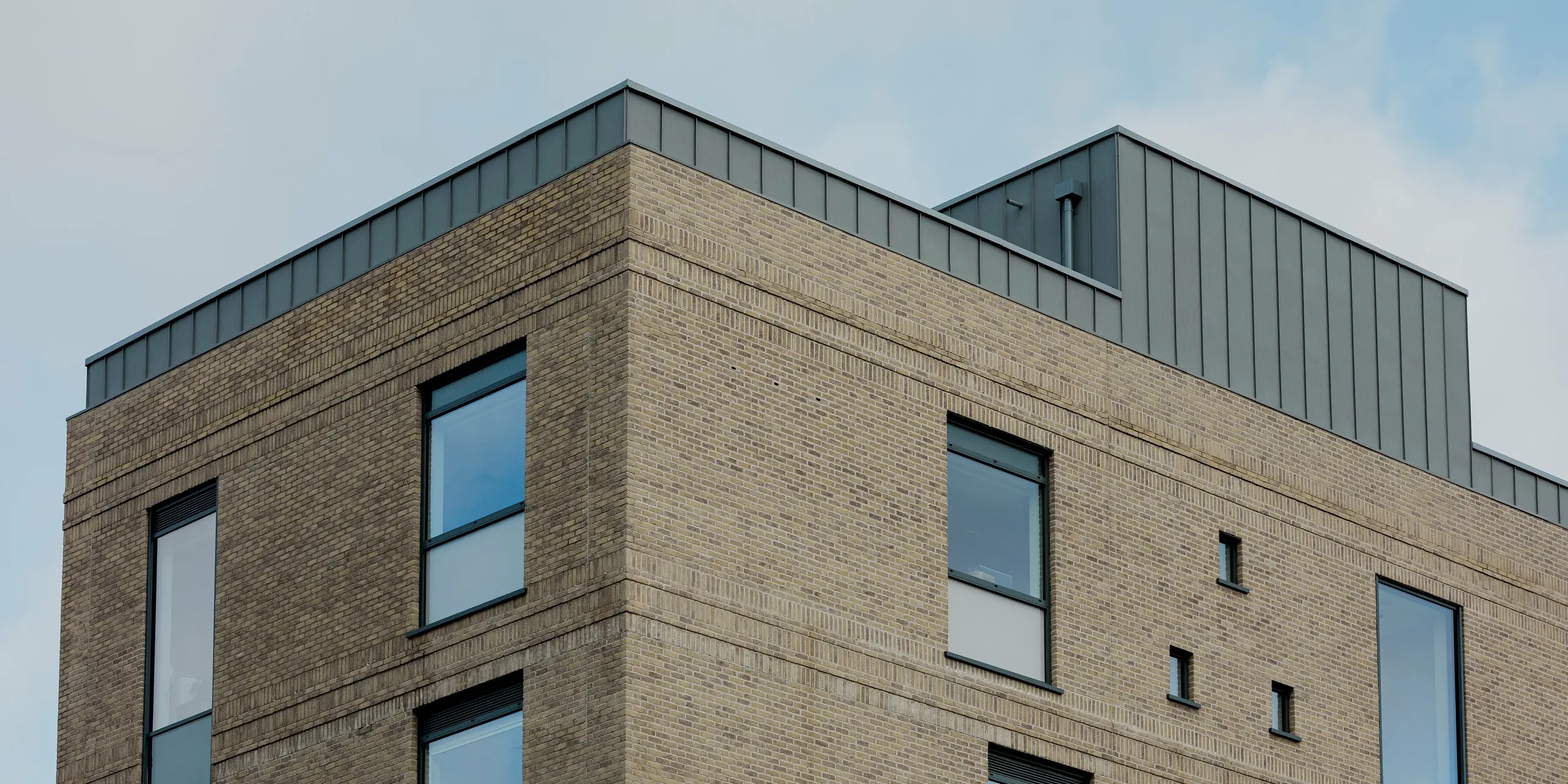 Die klare Gebäudeecke des Northern Regional College in Ballymena tritt mit hellem Klinkermauerwerk und präzisen Fensteröffnungen kraftvoll hervor. Entlang der Dachkante und am zurückgesetzten Aufbau bildet PREFALZ Aluminium in Patinagrau einen markanten, vertikal gegliederten Abschluss. Die Stehfalzbahnen setzen einen ruhigen metallischen Akzent über dem Mauerwerk und betonen die geometrische Architektur. PREFA Aluminium überzeugt durch Langlebigkeit, Witterungsbeständigkeit und eine zeitlose, hochwertige Lösung für moderne Bildungsbauten.