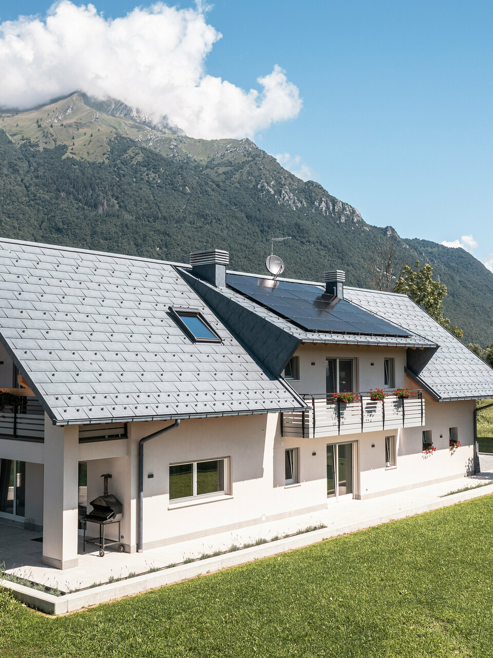 Drohnenaufnahme eines Hauses in Belluno, Italien, eingedeckt mit PREFA R.16 Dachplatten in P.10 anthrazit. Das Anwesen liegt in einer atemberaubenden Alpenlandschaft und kombiniert traditionelle Elemente wie freiliegende Holzgiebel mit modernen, langlebigen Baumaterialien, wodurch eine perfekte Symbiose aus Tradition und Moderne entsteht.