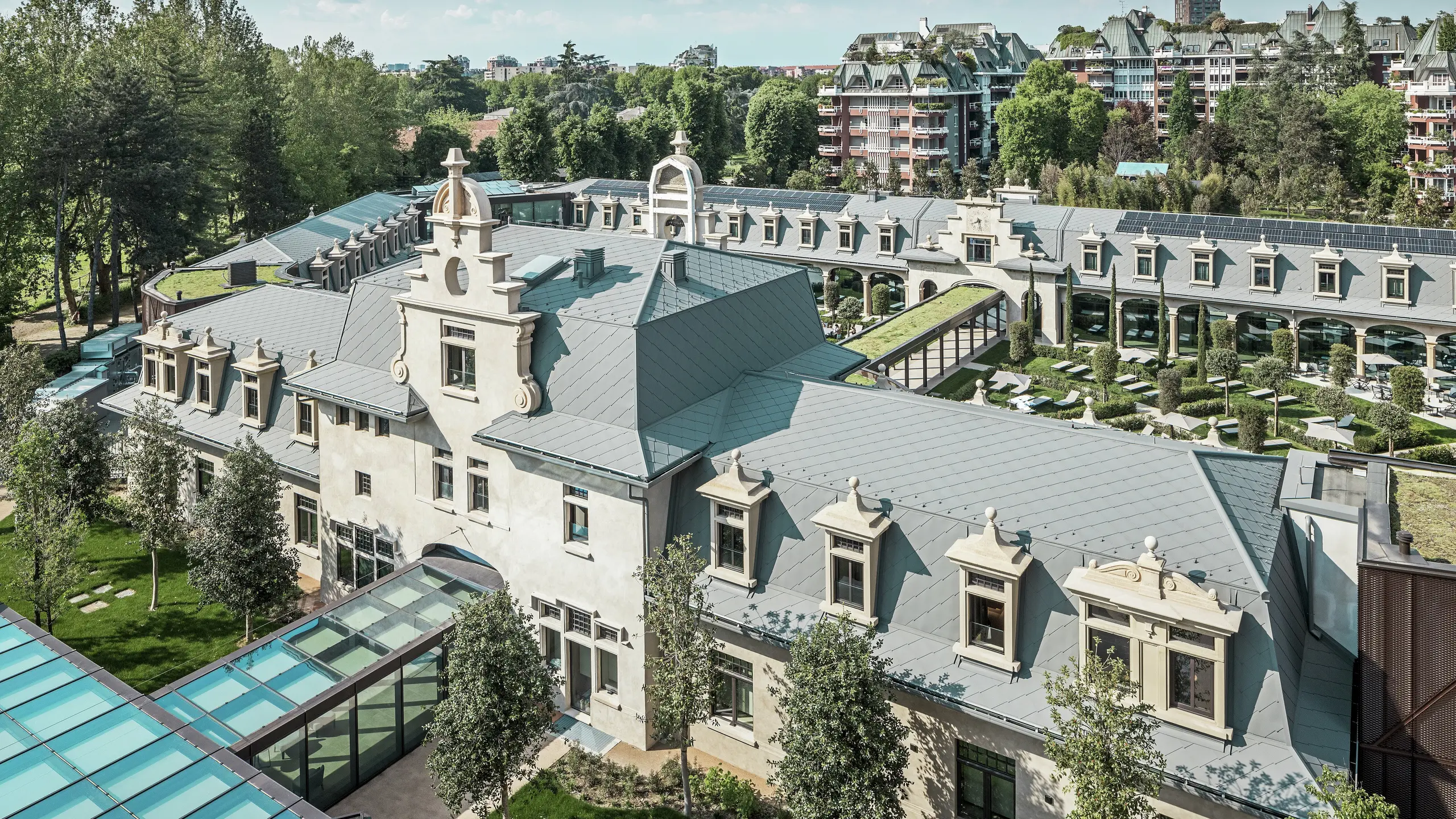 Gesamtansicht der imposanten Anlage „De Montel – Terme Milano“ mit denkmalgeschützter Jugendstilarchitektur und modernem Wellnessambiente. Die großflächige Dacheindeckung mit PREFA Dachrauten 44 × 44 in P.10 Hellgrau verleiht dem Komplex eine elegante Einheitlichkeit. Integrierte PV-Module, begrünte Dächer und Glasverbindungen zwischen den Baukörpern spiegeln den Anspruch an Nachhaltigkeit und zeitgemäßes Design im urbanen Kontext wider.