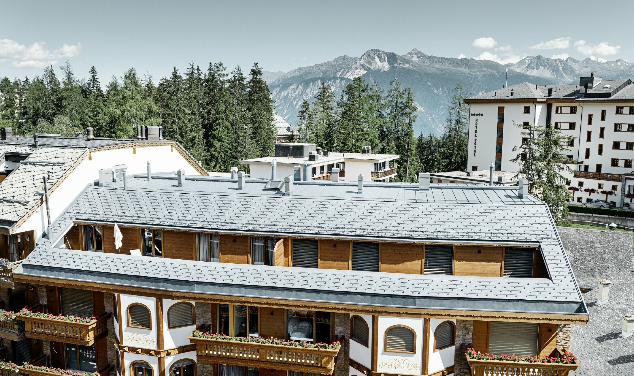 Mehrfamilienhaus in Crans-Montana mit Bergen im Hintergrund und einer Fassade mit verspielten Holzelementen mit einem PREFA Aluminium Dachschindel-Dach in steingrau 