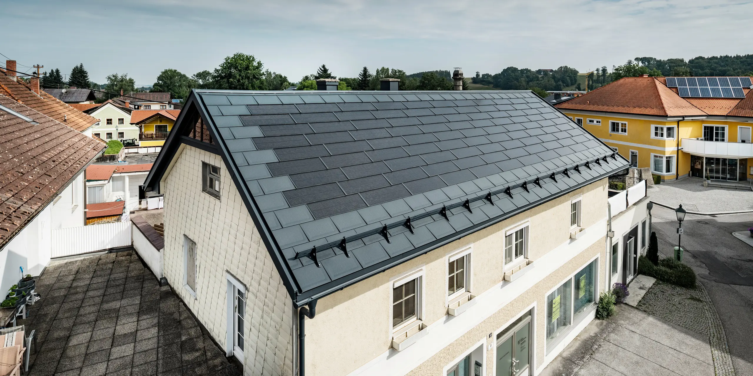 Utsikt ovenfra fra en enebolig i Mettmach i Østerrike. Den eldre bygningen ble renovert med antrasittfargede PREFA solcelle takplater. Snøbeskyttelsessystemene og de klare linjene på solcelle takplater understreker den rene utførelsen av PREFAs aluminiumsprodukter. Det oransjefargede tegltaket på nabobygningen danner en kontrast i bakgrunnen. Solcellemodulene er montert på taket av naboeiendommen i en klassisk på-tak-løsning, noe som viser mangfoldet i energieffektiv takdesign.