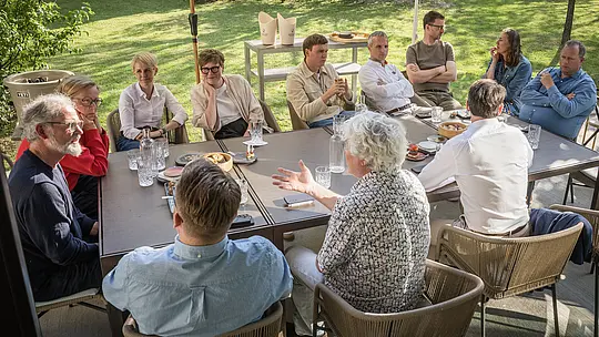PREFARENZEN 2027 Expertendialog im grünen Garten einer Winzerresidenz: Architekten und Designexperten sitzen um einen großen Tisch, diskutieren lebhaft und vertieft über moderne Architektur. Sonnenlicht fällt durch Bäume und sorgt für warme Akzente auf Gesichtern und Kleidung. Auf dem Tisch liegen Gläser, Wasserflaschen und kleine Häppchen, während im Hintergrund PREFA Aluminiumprodukte durch klare Linien und Langlebigkeit sichtbar werden. Die Szene zeigt, wie PREFA Dächer und Fassaden Design, Funktionalität und nachhaltige Ästhetik verbinden und kreative Diskussionen inspirieren.