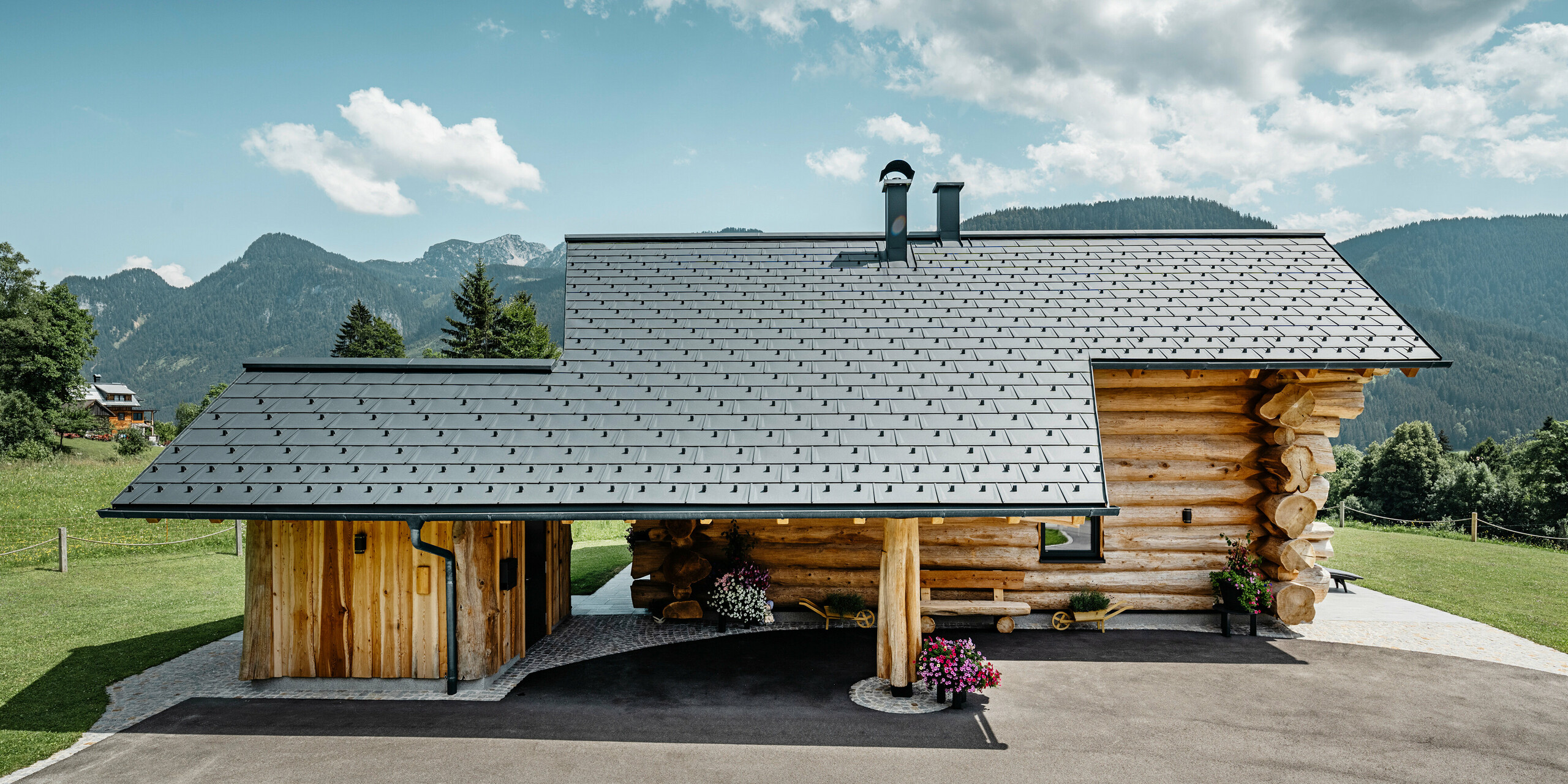 Seitenansicht eines einladenden Blockhauses in Gosau, Österreich, charakterisiert durch ein robustes PREFA Aluminiumdach mit Dachplatten R.16 in P.10 Anthrazit. Das Bild zeigt die harmonische Verbindung zwischen traditioneller Blockbauweise und moderner, hochwertiger Dacheindeckung, welche die Langlebigkeit und das ästhetische Design von PREFA Aluminiumprodukten widerspiegelt. Das Holzhaus steht vor einer Kulisse aus sanften grünen Hügeln und einem majestätischen Bergpanorama, was die naturnahe Umgebung unterstreicht.