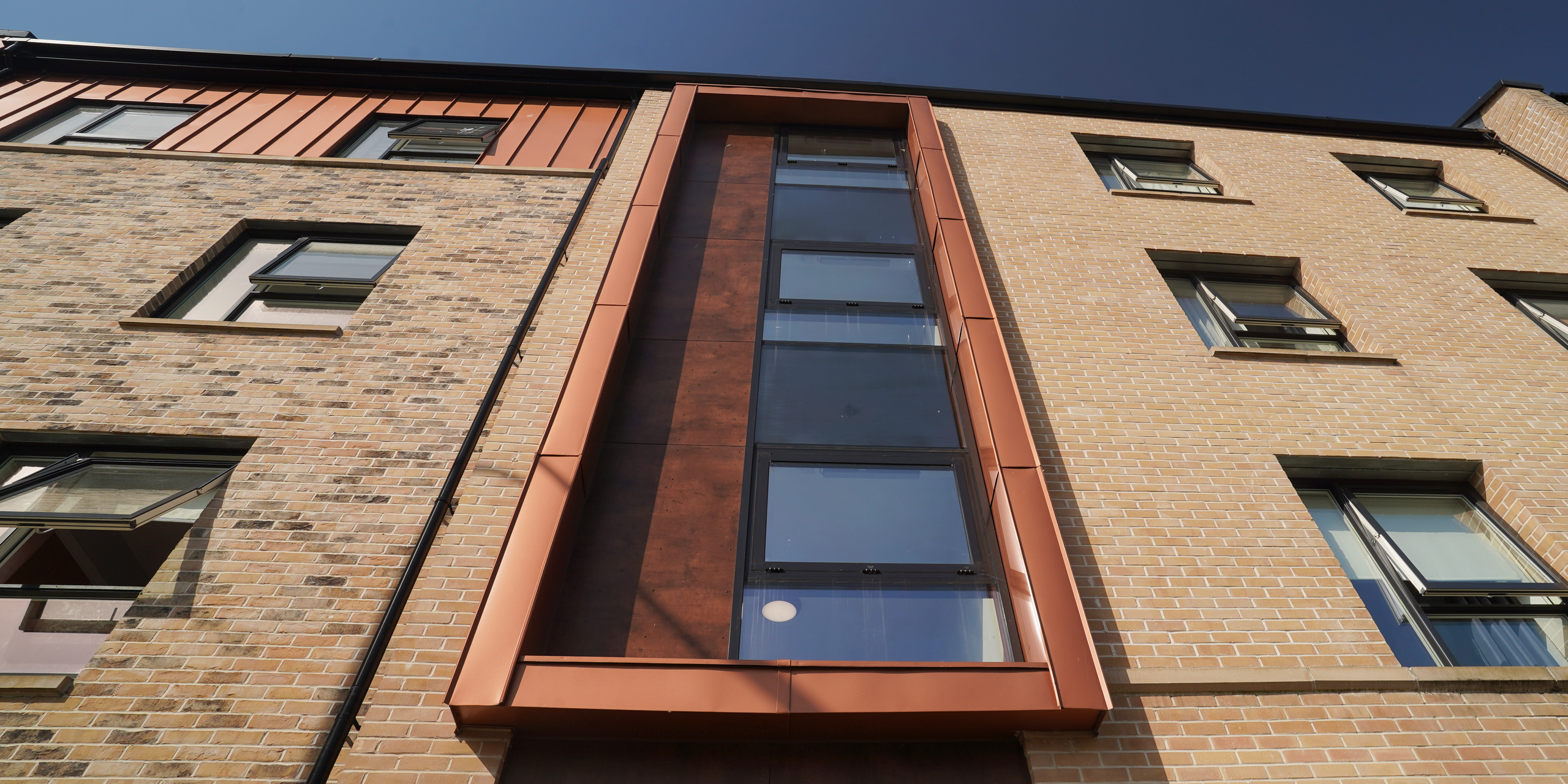 Detailansicht der Fassade eines Wohngebäudes in der Nethan Street in Glasgow, charakterisiert durch die Verkleidung mit FALZONAL in Neukupfer. Die kupferfarbene Verkleidung aus Aluminium rund um die Fensterpartien setzt moderne Akzente gegenüber der klassischen Ziegelsteinfassade und zeigt das Zusammenspiel von innovativen Materialien und traditionellem Design. Das hochwertige Aluminiumprodukt von PREFA unterstreicht die Ästhetik des Gebäudes und gewährleistet zugleich dessen Beständigkeit und Wetterfestigkeit.