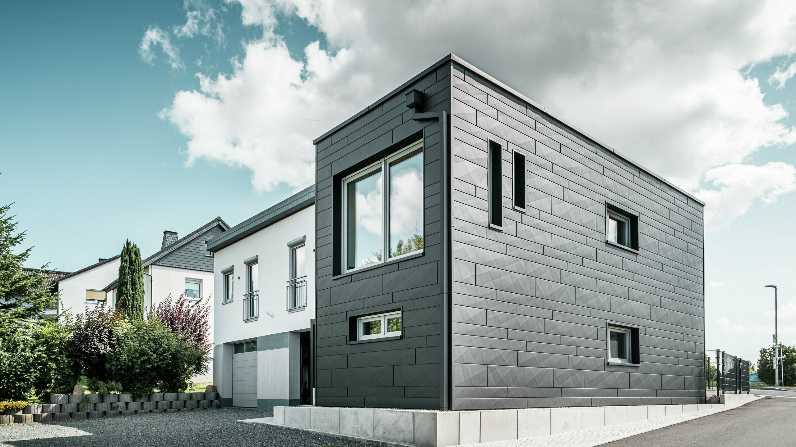 De stijlvolle en moderne gevel van de gerenoveerde eengezinswoning in Teuschnitz is voorzien van de duidelijk gestructureerde PREFA Siding.X-panelen in P.10 zwart. De geometrische opstelling van de hoogwaardige plaatpanelen geeft het gebouw een opvallende, tijdloze uitstraling. De ramen zijn omlijst met zwarte aluminium composietplaten, wat het ontwerp harmonieus afrondt. De geïntegreerde PREFA dakafwatering met vierkante buis zorgt voor een functionele en esthetische oplossing.