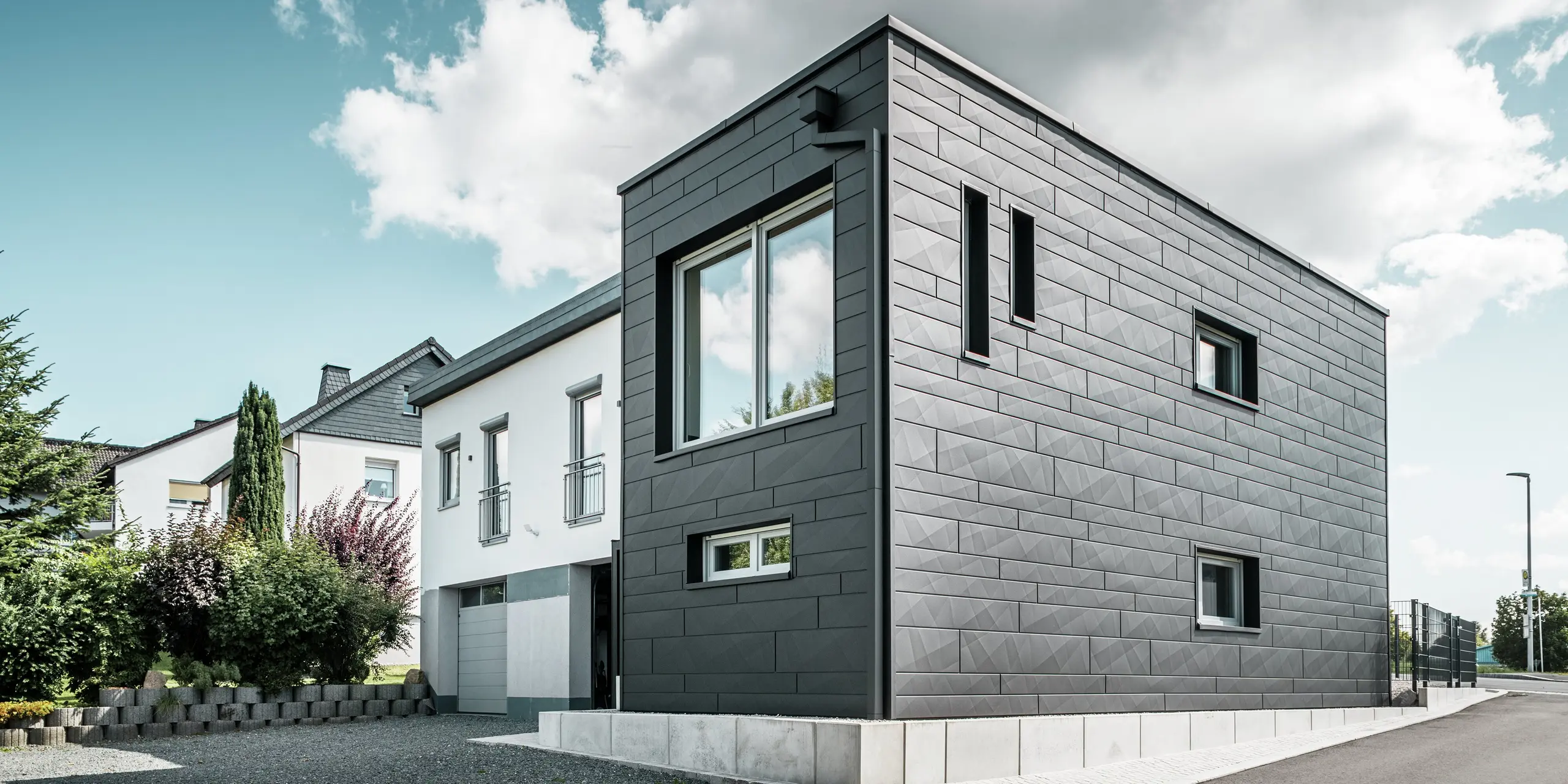 De stijlvolle en moderne gevel van de gerenoveerde eengezinswoning in Teuschnitz is voorzien van de duidelijk gestructureerde PREFA Siding.X-panelen in P.10 zwart. De geometrische opstelling van de hoogwaardige plaatpanelen geeft het gebouw een opvallende, tijdloze uitstraling. De ramen zijn omlijst met zwarte aluminium composietplaten, wat het ontwerp harmonieus afrondt. De geïntegreerde PREFA dakafwatering met vierkante buis zorgt voor een functionele en esthetische oplossing.