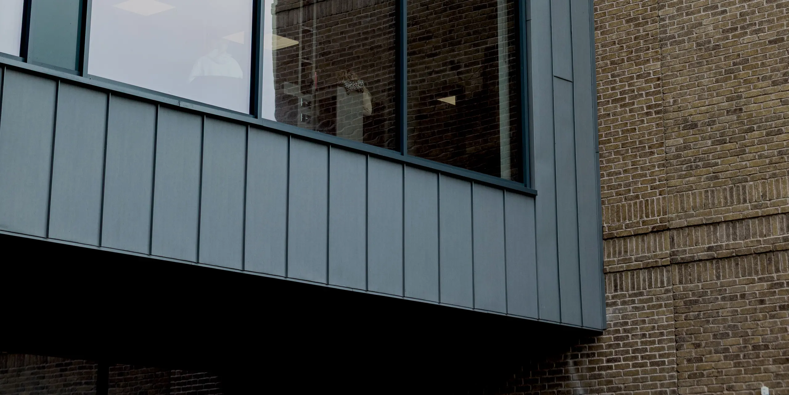 Ein markanter Fassadenausschnitt des Northern Regional College in Ballymena zeigt eine großzügige Glasfront, präzise eingefasst von PREFALZ Aluminium-Stehfalzbahnen in Patinagrau. Die vertikale Struktur des Aluminiums betont die klare Linienführung und rahmt den auskragenden Baukörper elegant ein. Im Kontrast dazu steht das fein strukturierte Klinkermauerwerk im Hintergrund. Die hochwertige PREFA Aluminiumfassade überzeugt durch Langlebigkeit, Witterungsbeständigkeit und eine ruhige, moderne Optik für zeitgemäße Bildungsarchitektur.