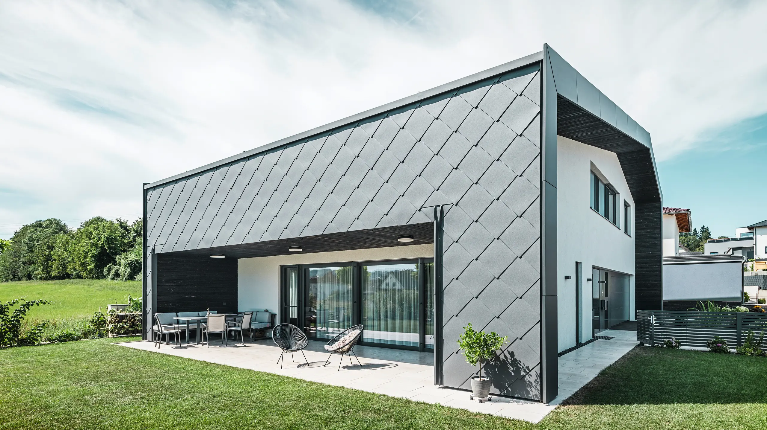 Velstelt hageområde til en enebolig i Aschach. Bygningen er stilfullt kledd med mørk grå PREFA aluminium veggrombe 44 × 44. Den overbygde terrassen gir et koselig sitteplass med utsikt over den frodige grønne plenen. Den elegante aluminiumsbekledningen i kombinasjon med hvite pussflater gir en moderne og værbestandig arkitektur. Store vindusflater slipper mye dagslys inn i rommene og skaper en åpen, lys atmosfære.