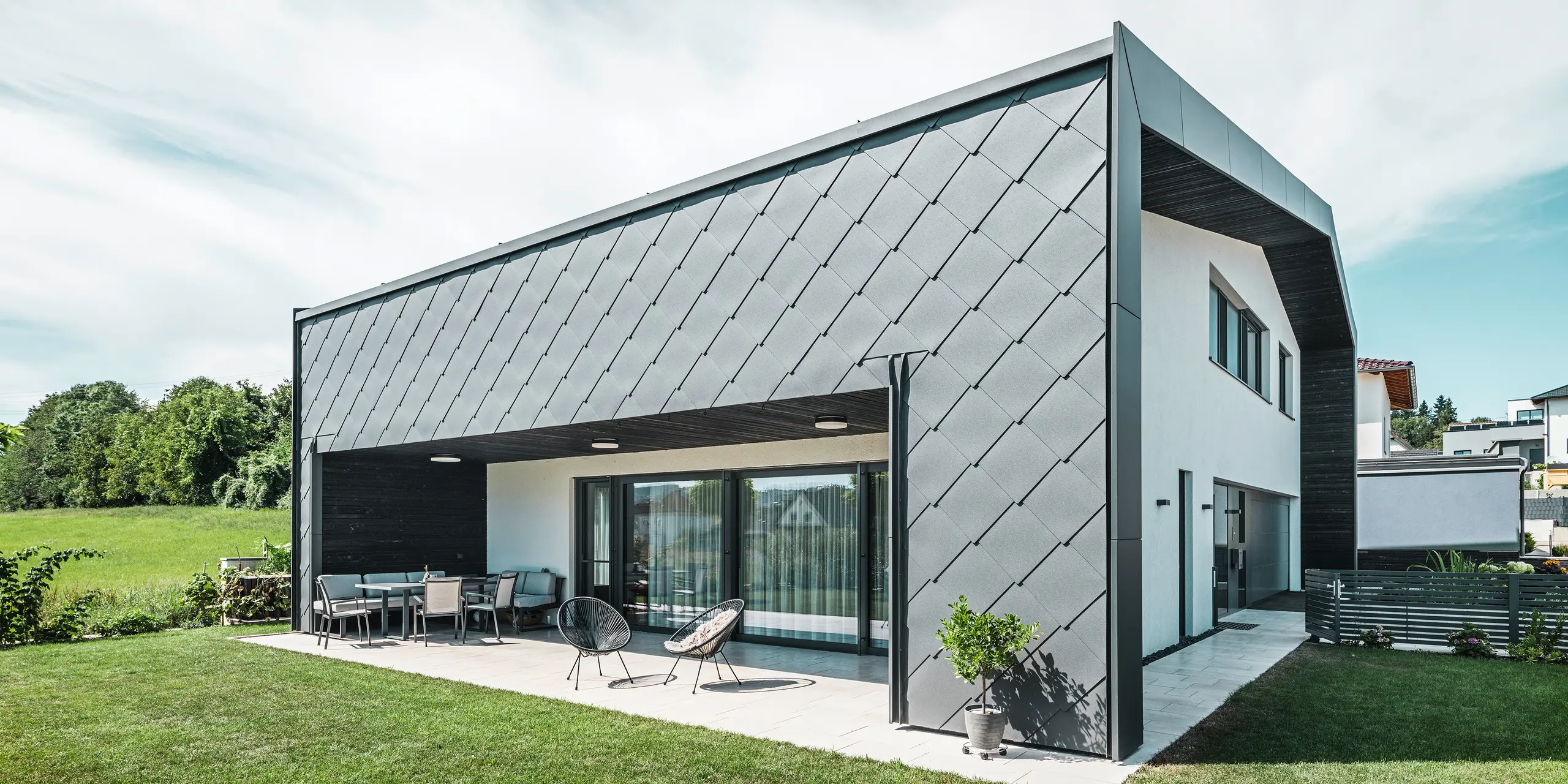 Velstelt hageområde til en enebolig i Aschach. Bygningen er stilfullt kledd med mørk grå PREFA aluminium veggrombe 44 × 44. Den overbygde terrassen gir et koselig sitteplass med utsikt over den frodige grønne plenen. Den elegante aluminiumsbekledningen i kombinasjon med hvite pussflater gir en moderne og værbestandig arkitektur. Store vindusflater slipper mye dagslys inn i rommene og skaper en åpen, lys atmosfære.