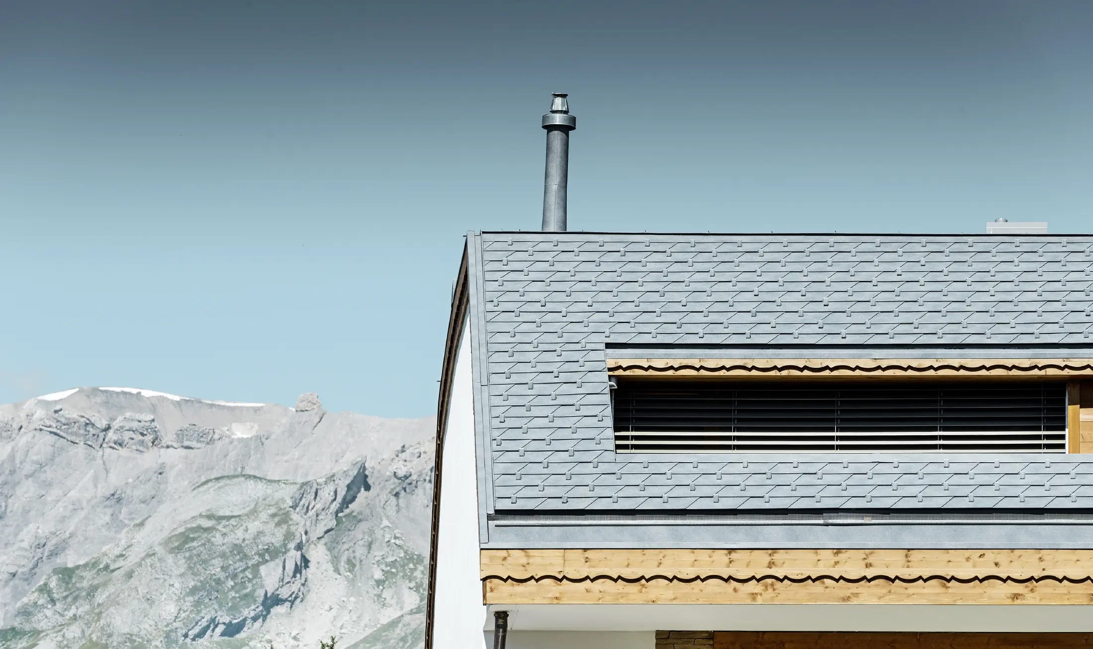 Mehrfamilienhaus in Crans-Montana mit Bergen im Hintergrund und einer Fassade mit verspielten Holzelementen mit einem PREFA Aluminium Dachschindel-Dach in steingrau 