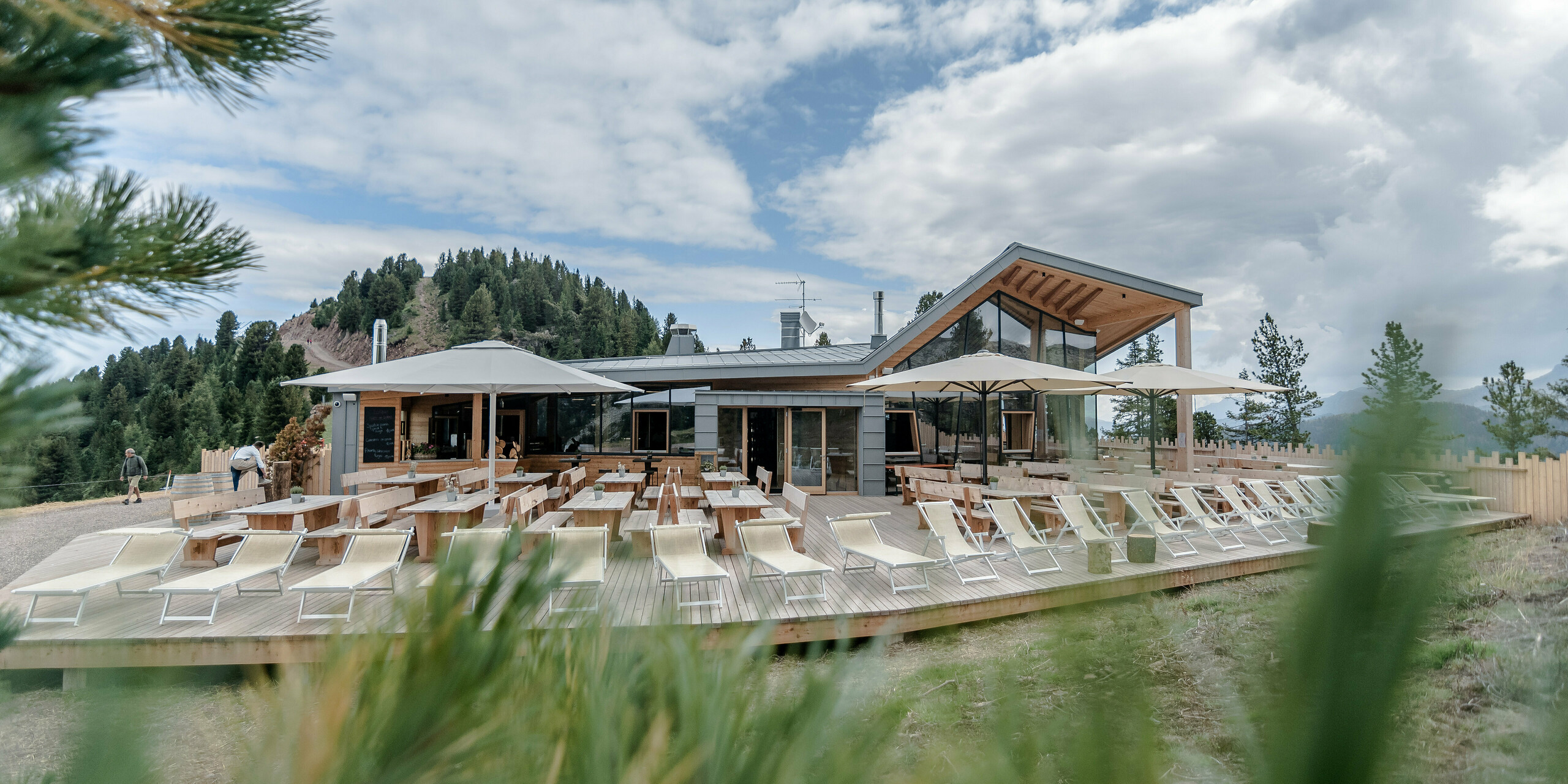 Die Außenterrasse des Ristorante Busabelle in Italien bietet eine großzügige und einladende Atmosphäre mit zahlreichen Sonnenliegen und Holztischen, die von eleganten Sonnenschirmen beschattet werden. Der Hauptbau des Restaurants, charakterisiert durch eine moderne Architektur mit hellgrauer PREFALZ Fassade und Holzelementen, fügt sich nahtlos in die bewaldete Berglandschaft im Hintergrund ein.