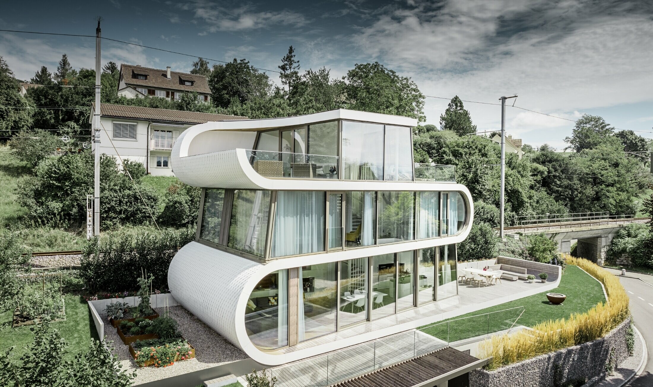 Svært moderne enebolig tegnet av arkitekt Camenzind fra Zürich. Et buet bånd forbinder de enkelte flatene med hverandre. De runde stedene ble kledd med små PREFA takromber i helt hvitt. Huset har mange og store glassflater slik at det virker å være helt åpent.