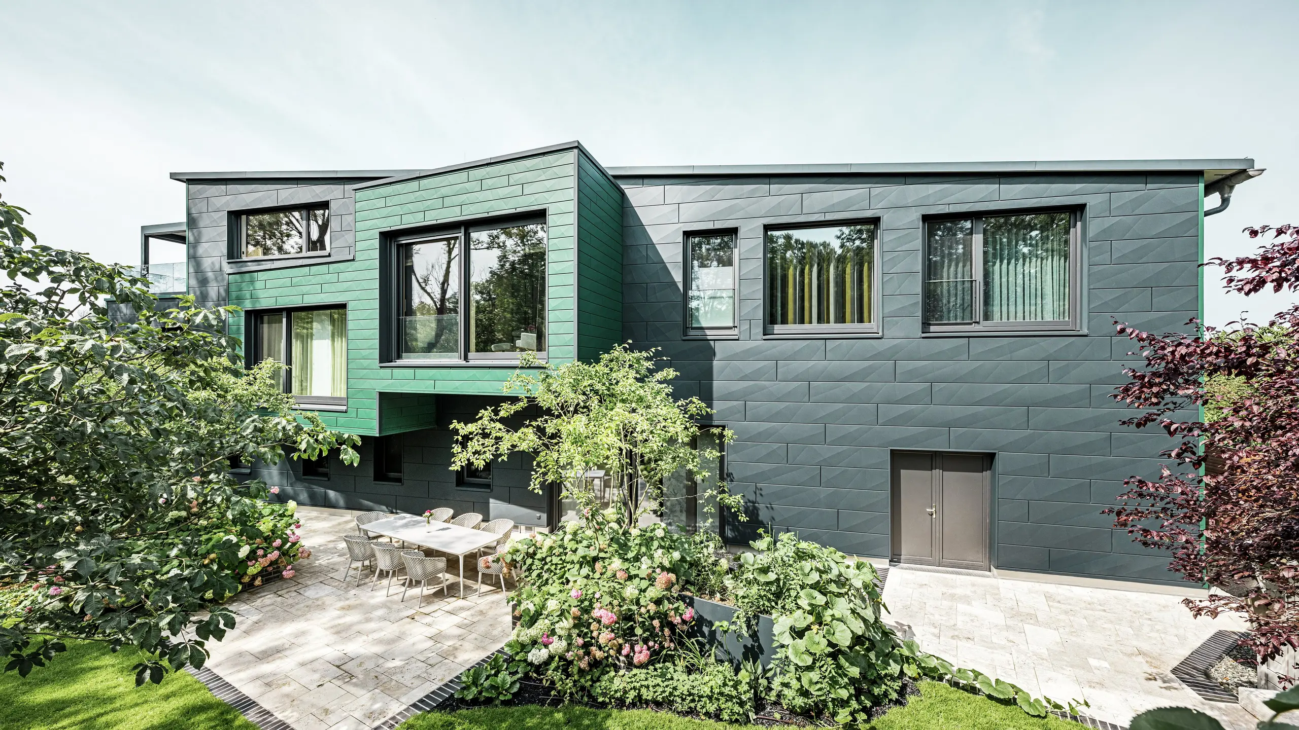 Enkeltboligen i Türkheim imponerer med sin ekstraordinære fasade, som er kledd med PREFA Siding X-paneler i fargene antrasitt og mosegrønn. De klare linjene og det markante fargevalget på aluminiumpanelene gir bygningen et moderne og elegant utseende. I forgrunnen ser man den romslige hagen med en stilfull terrasse og et spisebord med moderne stoler. Fargerike blomsterbed og sunne planter skaper en levende, grønn kontrast til den robuste fasaden. Den holdbare og vedlikeholdsfrie aluminiumsfasaden beskytter bygningen og sørger for tidløs skjønnhet.