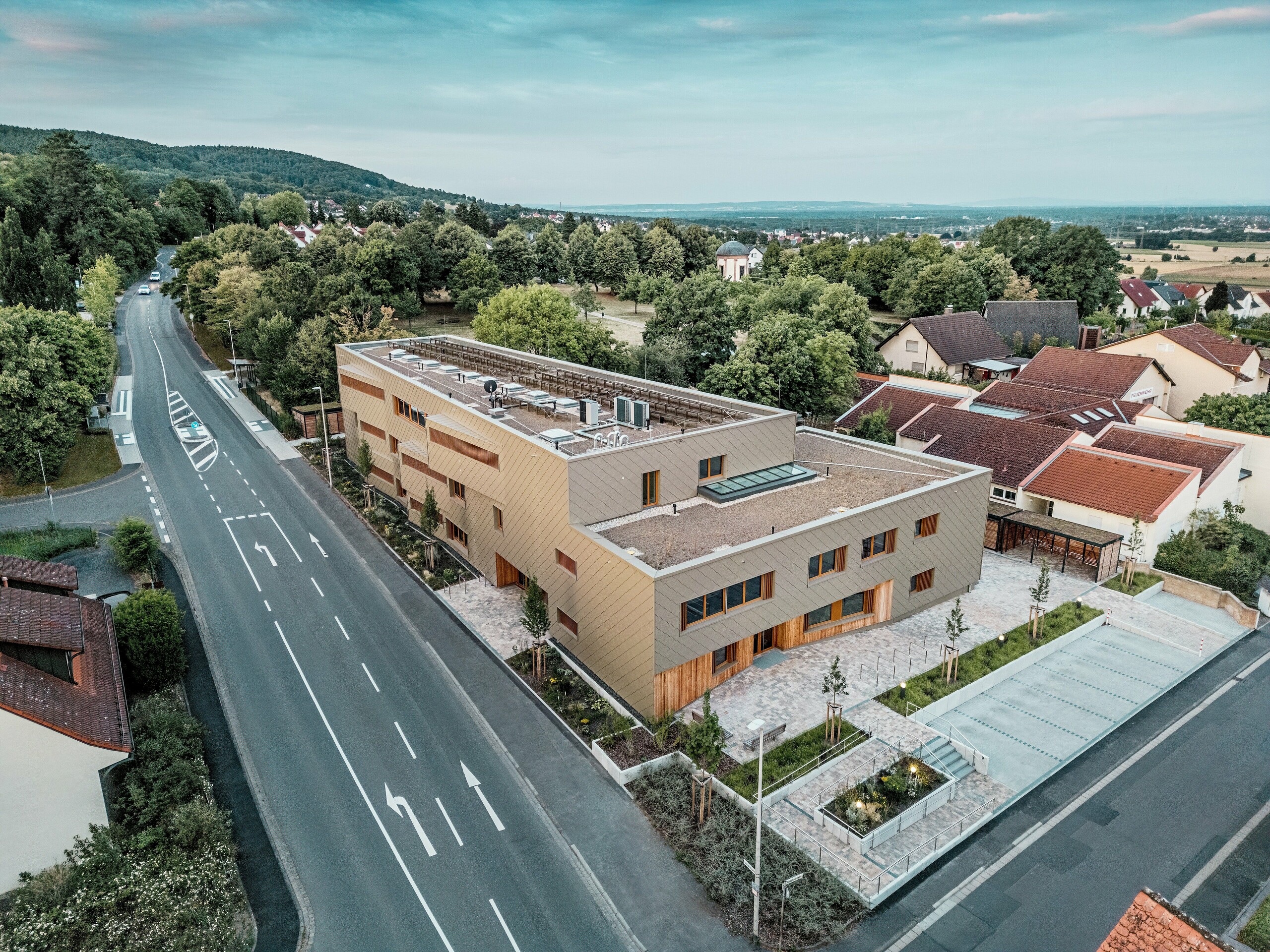 Die eindrucksvolle Gesamtgestaltung der kommunalen Kindertagesstätte in Alzenau wird auf der Luftaufnahme sichtbar. Das winkelförmige Hybridgebäude ist harmonisch in ein ruhiges Wohnviertel am Rande des Spessarts eingebettet. Die Fassade aus bronzefarbenen PREFA Wandrauten 44 × 44 verleiht dem Baukörper eine markante, hochwertige Optik und betont die moderne Architektur. Begrünte Dachflächen, großzügige Außenanlagen und eine klare Verkehrsführung unterstreichen das nachhaltige Gesamtkonzept. Die Kombination aus langlebigem Aluminium, Holz und Recyclingbeton erfüllt höchste ökologische Standards. Diese Perspektive hebt die durchdachte städtebauliche Integration und den multifunktionalen Charakter des Gebäudes hervor – ein zukunftsweisendes Projekt für Bildung, Wohnen und Nachhaltigkeit.