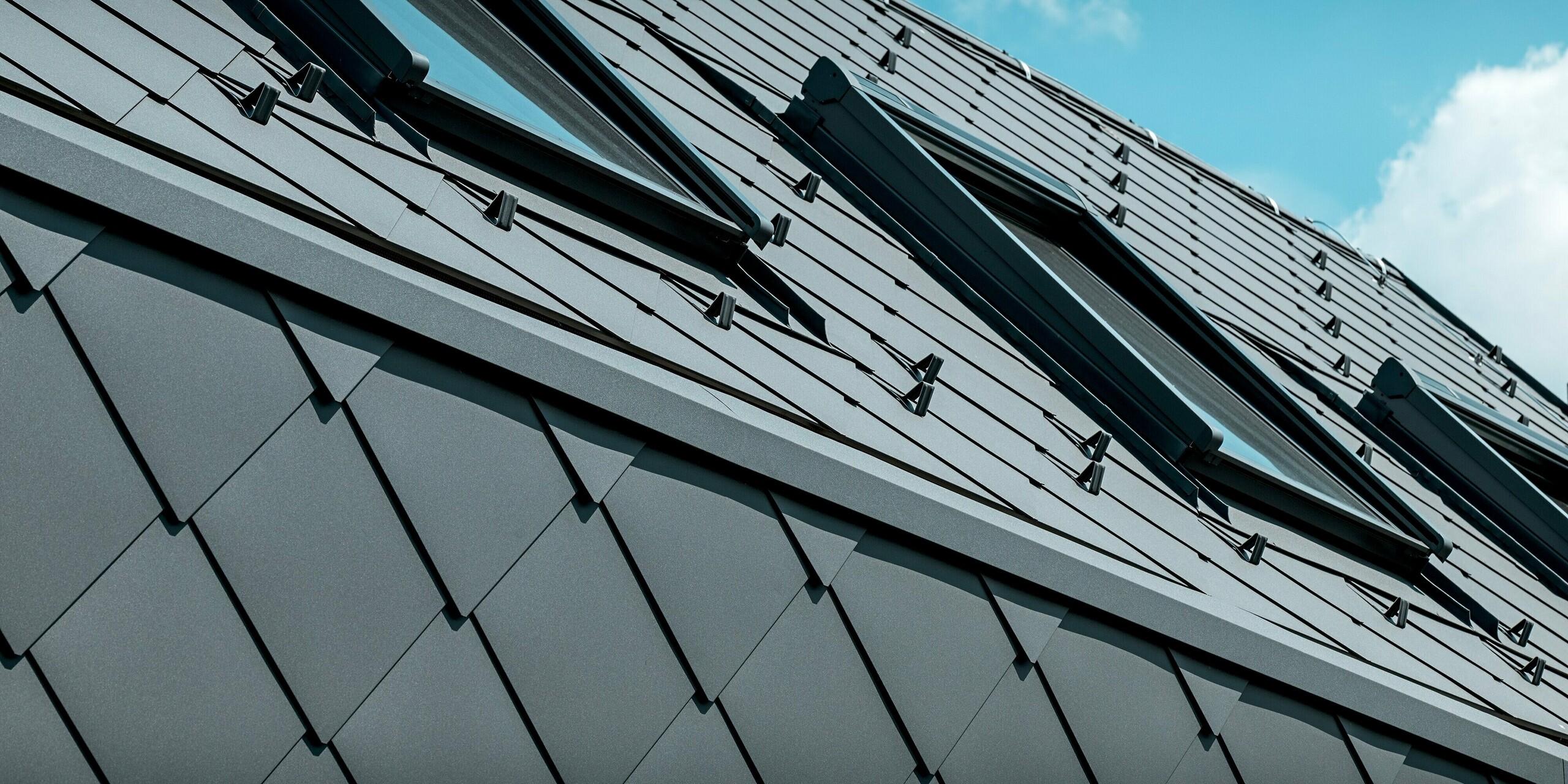 Das Bild zeigt eine dynamische Detailaufnahme des Dachs des Einfamilienhauses in Trnava‑Modranka, eingedeckt mit PREFA Dachrauten 44 × 44 in P.10 Dunkelgrau. Mehrere Dachfenster setzen moderne Akzente und brechen die gleichmäßige Aluminiumstruktur auf elegante Weise. Die saubere Verlegung, integrierten Schneestopper und die widerstandsfähige Oberfläche unterstreichen die hohe Qualität der PREFA Produkte. Die langlebige Aluminiumdeckung bietet optimalen Witterungsschutz, Formstabilität und eine markante, moderne Optik im Zusammenspiel mit dem klaren Himmel.